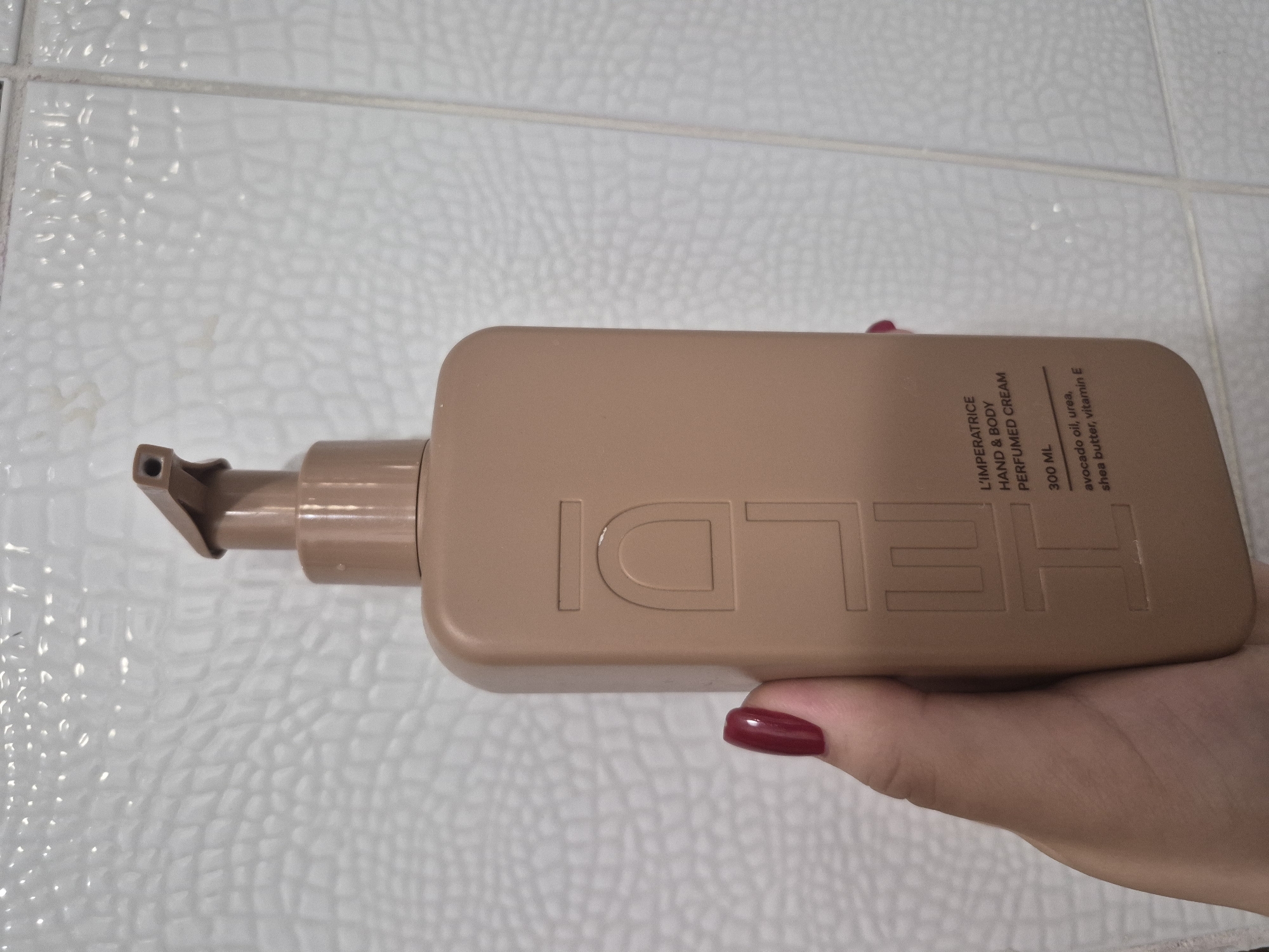 A person's hand holds a light brown, rectangular cosmetic bottle with a pump dispenser against a white tiled background. The hand, likely female, has dark red nail polish on the fingernails. The bottle is embossed with the large letters "HELDI". Vertically printed text on the bottle identifies it as "L'IMPERATRICE HAND & BODY PERFUMED CREAM", containing "300 ML". Further details list key ingredients: "avocado oil, urea, shea butter, vitamin E". The background features white, square ceramic tiles with visible grout lines, suggesting an indoor setting such as a bathroom or kitchen. The lighting is uniform, indicating artificial light. The image captures a casual display or use of the product within a domestic environment in Tobolsk, Russia.