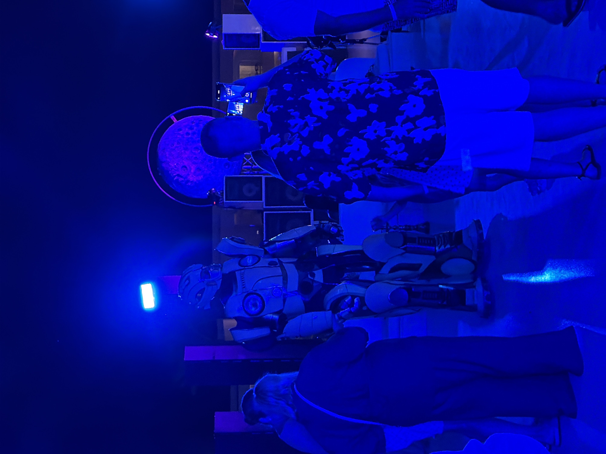 In Sharm El Sheikh, Egypt, a tall, yellow and black robot costume stands center frame, illuminated by blue lights. Three people are visible in the foreground: two women on the left facing the robot, one in a dark outfit and the other in a dark top and what appear to be light pants, and a man on the right facing away from the robot, holding up a phone to take a picture. He is wearing a dark, floral-patterned shirt and white shorts. Behind the man, speakers are stacked, and a large, circular, purple-lit object resembling a planet or moon is visible. The scene appears to be an outdoor event at night, likely a party or performance, given the lighting and music equipment.