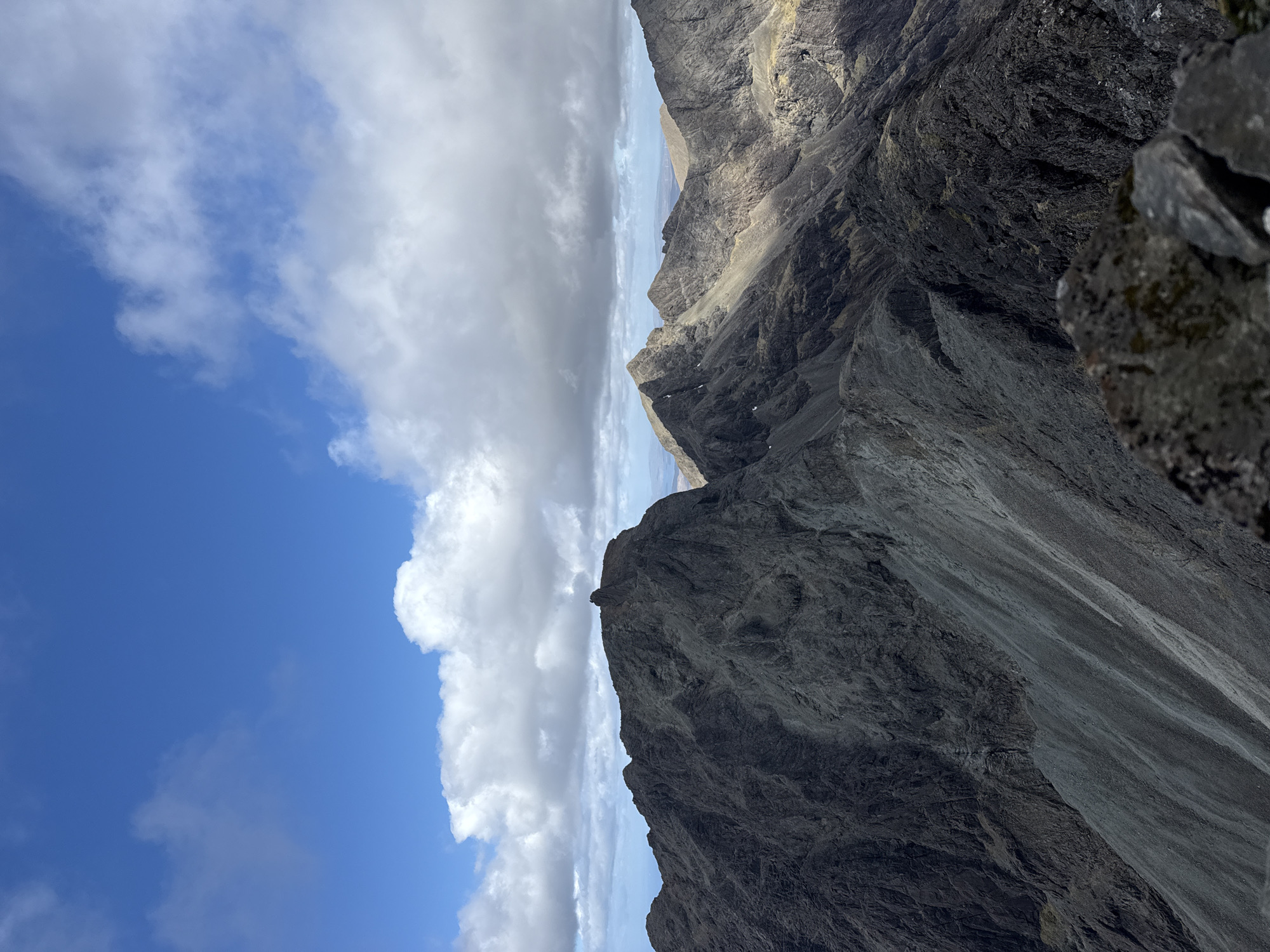 The image depicts a mountainous landscape under a partly cloudy sky. The mountains are steep and rocky, with varying shades of gray and brown suggesting different rock types or lighting. The sky is divided between patches of clear blue and dense, white and gray clouds. The perspective suggests the photographer is at a high vantage point. The location is unknown.