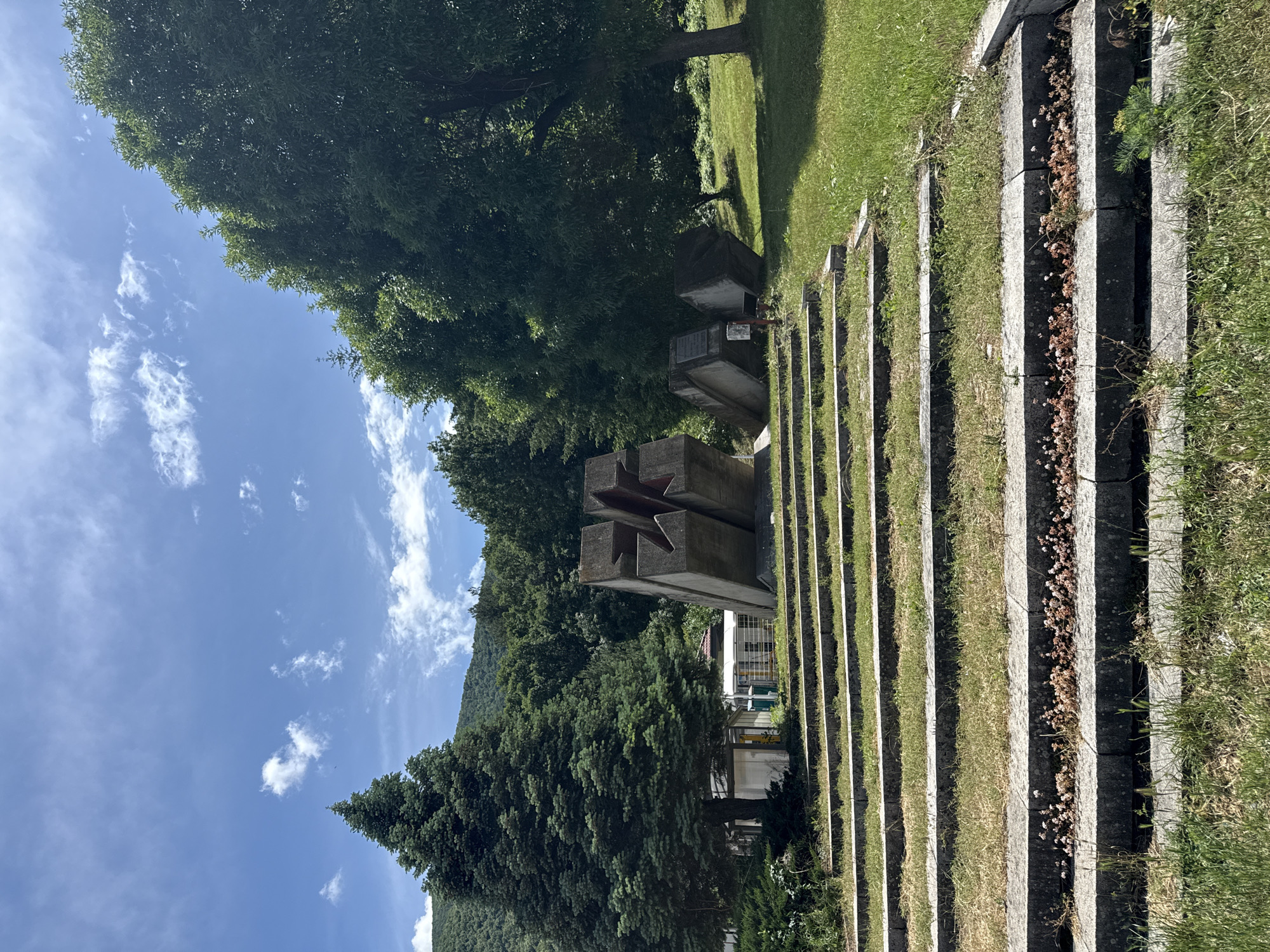 This image captures an outdoor memorial complex or cemetery in Vetren Dol, Bulgaria, under a clear, sunny sky. The scene is static, devoid of people or ongoing activity.
In the midground, a prominent brutalist-style concrete monument stands out, featuring a large, deeply carved five-pointed star on its front face. This central monument is flanked on its right by a series of concrete steps or terraces that extend into the foreground. These steps are interspersed with strips of green grass, some appearing well-maintained, while others contain dried, light brown foliage.
Behind the main star monument, several other rectangular concrete structures are visible, likely grave markers or smaller memorial stones. One of these smaller structures appears to have engraved text, possibly in Cyrillic, with discernible numbers that look like "194...", suggesting a historical date.
The background is dominated by a dense array of green trees, including both broadleaf and coniferous varieties, indicating a forested area. Beyond these trees, a mountain range, also covered in forest, rises under a bright blue sky scattered with fluffy white cumulus clouds. The clear skies and visible shadows suggest it is daytime, likely morning or early afternoon. The overall setting conveys a sense of solemnity and historical remembrance, typical of war memorials or significant commemorative sites.