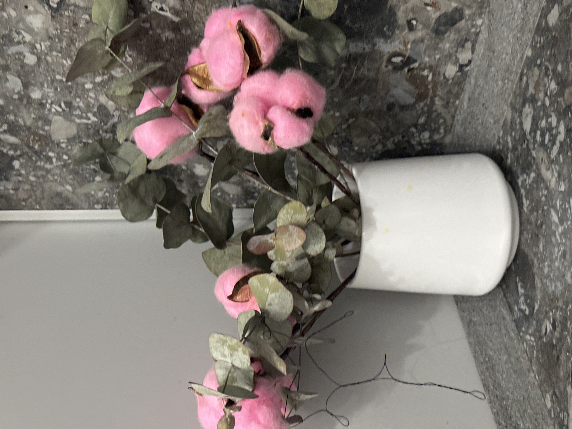 A close-up shot captures a white ceramic vase filled with dried eucalyptus branches adorned with bright pink cotton bolls. The vase rests on a speckled gray surface, possibly a countertop or shelf, adjacent to a wall with a textured, dark gray and white pattern. The background is blurred, but the overall impression is one of a decorative arrangement. The image does not provide information about the city of Krasnodar, Russia.