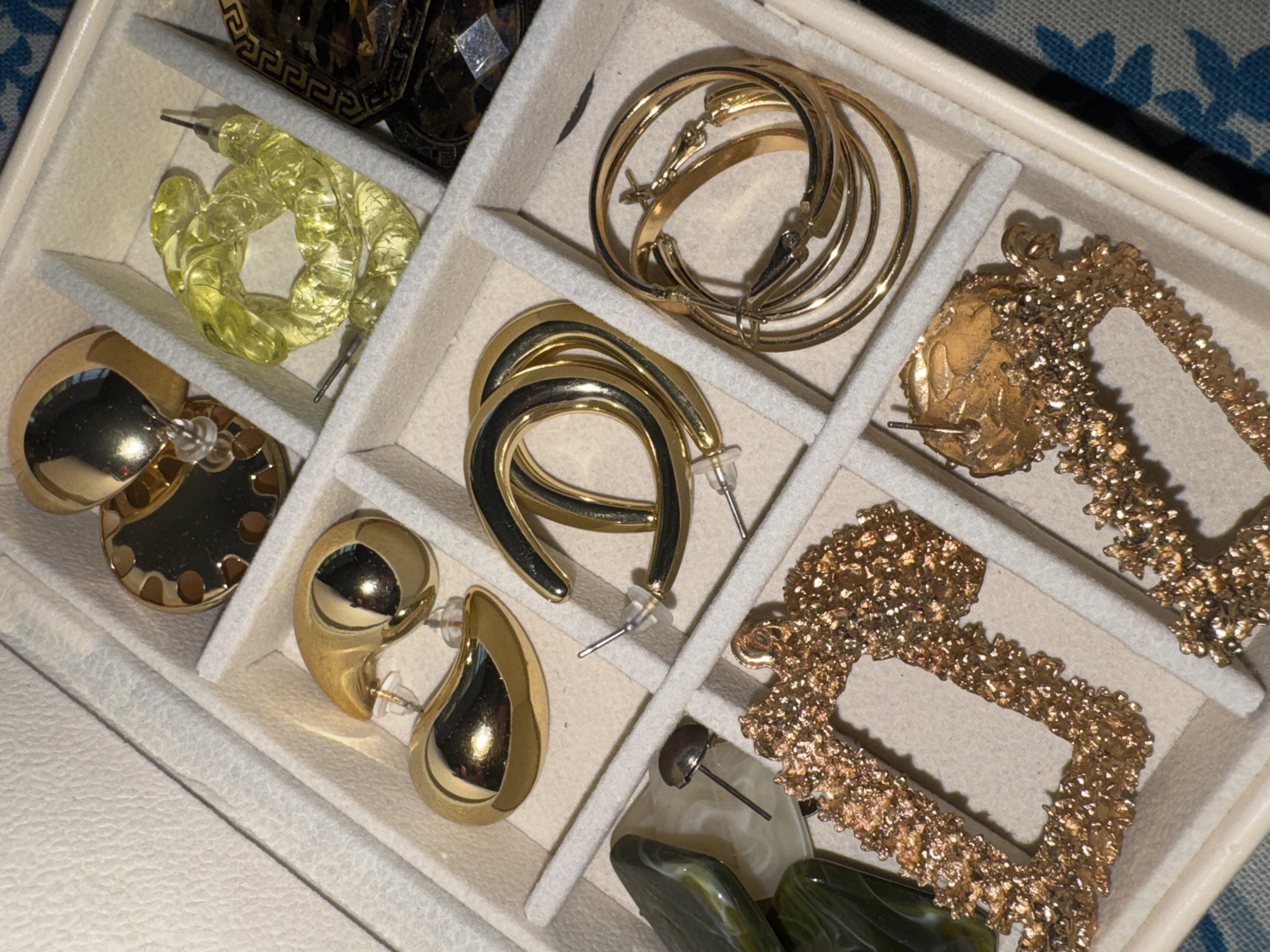 The image shows a top-down view of an open jewelry box, filled with various earrings. The box is divided into compartments, with each holding a different pair or set of earrings. In the top left compartment, a pair of translucent yellow, twisted hoop earrings are visible. Next to them, in the top middle compartment, several pairs of gold hoop earrings are stacked. The top right compartment contains a single gold earring with a textured, circular face.
In the middle row, the left compartment holds a pair of large, shiny gold hoop earrings with cut-out details. To their right, a pair of gold, c-shaped hoop earrings are displayed. The middle right compartment contains another gold earring with a textured, rectangular shape.
The bottom row shows a pair of polished gold, teardrop-shaped earrings in the left compartment. In the middle compartment of the bottom row, a pair of earrings with polished grey and green marbled, irregular shaped stones are visible, with one earring's post sticking out. To the right of this, in the bottom right compartment, another textured gold rectangular earring is present.
The overall setting appears to be indoors, with a blue and white patterned fabric visible in the background, suggesting the jewelry box is placed on a surface with this fabric. The lighting suggests it could be daytime, or well-lit indoors, as the metallic jewelry reflects light. No people or specific activities are depicted, only a collection of jewelry. There is no visible text in the image.