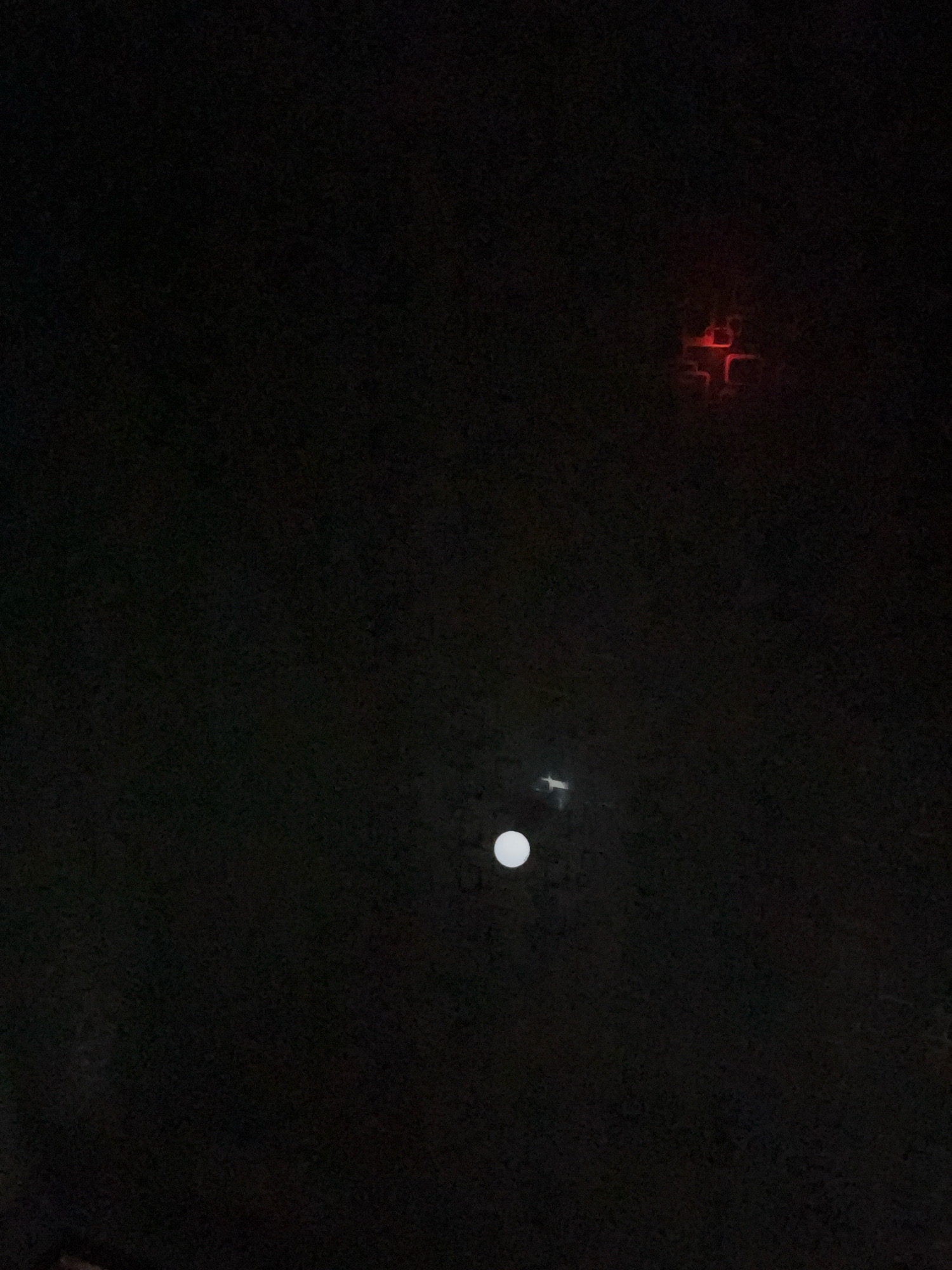 The image is a dark, slightly grainy photograph taken at night in Matesih, Indonesia. In the upper right quadrant, a faint red light source with a segmented, possibly geometric, pattern is visible. In the lower center of the frame, a small, bright white circular object, likely the moon, is present, with a faint white light object slightly above and to its left. The background is predominantly black, suggesting a clear night sky with minimal ambient light. There are no discernible people or animals in the image. The overall impression is a nocturnal scene, possibly urban or semi-urban, given the presence of artificial lights.