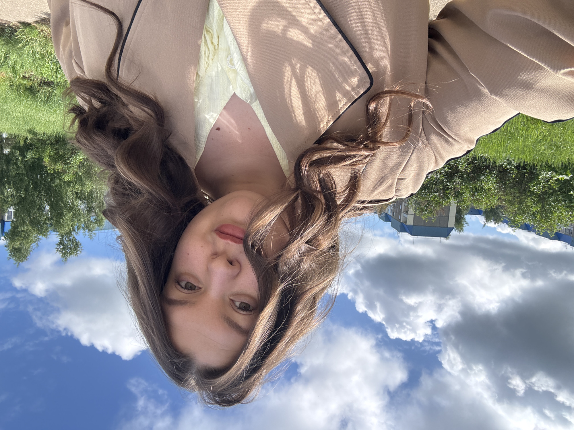 The image is a selfie of a young woman with long, wavy brown hair. She is wearing a beige blazer with black piping and a pale yellow top underneath. She has fair skin, brown eyes, and light pink lipstick.
The background is a mix of natural and urban elements. The sky is visible with blue patches and white clouds. Trees and greenery are on the left, suggesting a park or green space. In the distance, there's a building, possibly a residential apartment block, consistent with an urban setting in Russia, specifically Марковский.
The image is taken from a slightly elevated angle, looking down at the woman. The overall scene suggests a sunny day in a city park, possibly near residential areas.