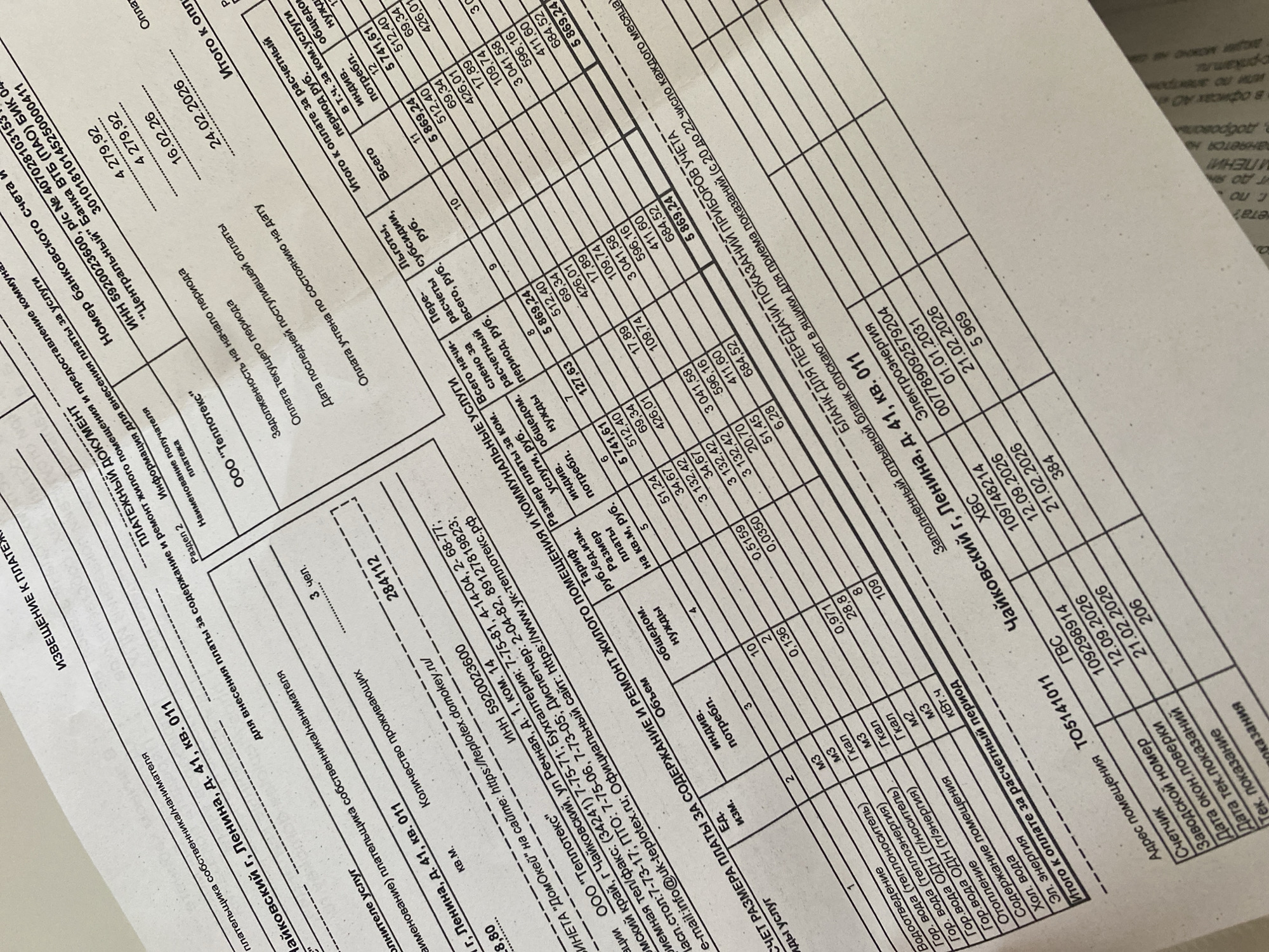 The image displays a physical paper document, identified as a payment notice and payment document, positioned on a light, neutral-colored surface. The document is titled "ИЗВЕЩЕНИЕ К ПЛАТЕЖУ" (Notice to Payment) and "ПЛАТЕЖНЫЙ ДОКУМЕНТ" (Payment Document). It details utility service charges for a residential property located at Chaikovsky city, Lenin Street, building 41, apartment 011 in Russia.
The document indicates "3 чел." (3 persons) as the number of residents. The service provider is "ООО "Теплотекс"" (LLC "Teplotex"), with its INN 5920023600 and contact details printed. The bill itemizes charges for various communal services, including water supply, hot water, heating, cold water, and electricity, along with maintenance and repair of the residential premises. Individual consumption, general household needs, and associated costs in Rubles are listed.
Key financial details include a total amount due of 5,869.24 Rubles ("Итого к оплате"), with a payment due date of 24.02.2026. A date of last received payment is noted as 16.02.2026. Sections for meter readings ("Счетчик"), serial numbers ("Заводской номер"), dates of last verification ("Дата окон. поверки"), and dates of current readings ("Дата тек. показаний") are present for various meters (e.g., GVS, HVS, Электроэнергия). An instruction to use a detachable form for submitting meter readings is also visible. The document is slightly angled and appears to be in good condition, without folds or tears. No people, actions, or interactions are visible in the scene.