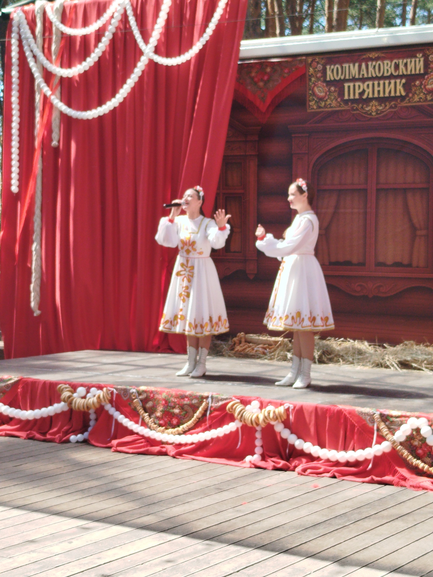 The image captures a scene on an outdoor stage in Zavodoukovsk, Russia, likely during a cultural festival. Two young women in traditional white embroidered dresses with red trim and floral headbands are performing. The woman on the left is holding a microphone and singing, while the woman on the right gestures with her hands. The stage is decorated with a red draped fabric adorned with strings of white beaded garlands and traditional baked goods resembling bagels. Behind the performers is a wooden backdrop painted to resemble a traditional Russian house with ornate window frames and a sign at the top reading "КОЛМАКОВСКИЙ ПРЯНИК" (Kolmakovsky Pryanik), which translates to "Kolmakovsky Gingerbread". The overall atmosphere suggests a folk performance or a celebration.