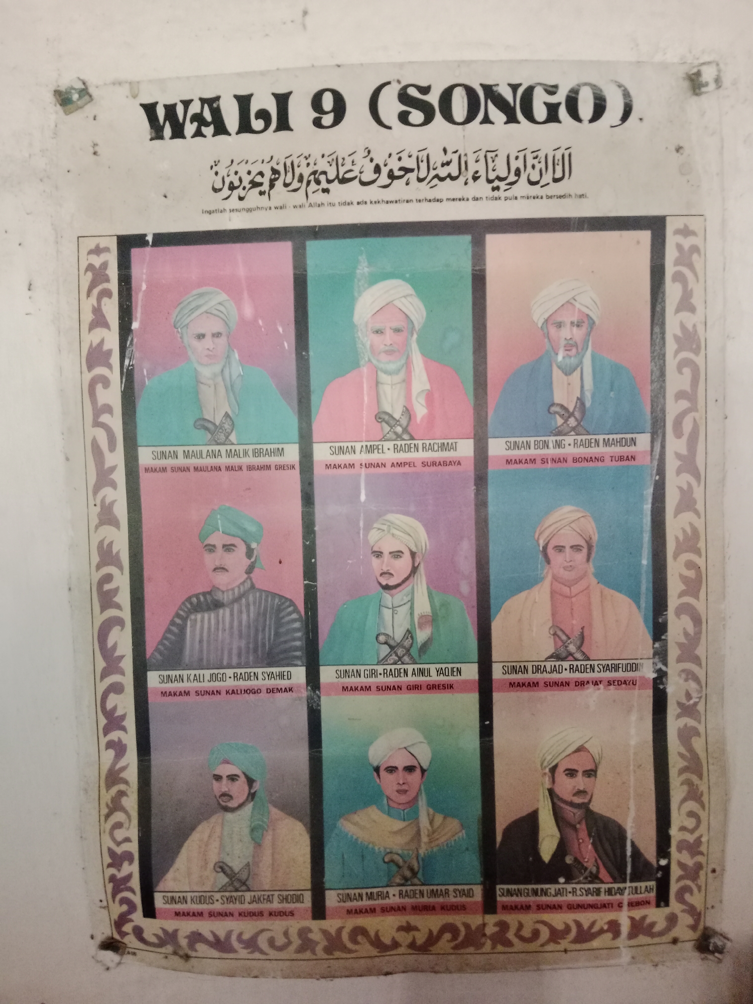 This is a photograph of a poster depicting the nine Walisongo, revered Sufi saints in Indonesia. The poster is mounted on a white wall, which shows signs of age and possibly some dampness or dirt. The text at the top of the poster reads "WALI 9 (SONGO)" in large black letters, with Arabic script below it and smaller Indonesian text that translates to "Remember, indeed the walis, Allah, there is no fear upon them, nor do they grieve."

The poster is divided into nine sections, each featuring a portrait of one of the Walisongo. Each portrait is accompanied by the name of the Wali and the location of their tomb. The individuals are depicted wearing traditional Islamic attire, including turbans and robes, and most are shown holding a dagger or sword. The artistic style of the portraits is illustrative and somewhat stylized, with vibrant, flat colors used for the backgrounds of each section.

The specific location in the city of Ringinrejo, Indonesia, is not explicitly visible in the photograph. However, the presence of the Walisongo poster suggests a context related to Islamic history and culture in Indonesia, potentially a religious school, mosque, or community center in Ringinrejo. The overall scene conveys a sense of historical reverence and cultural significance.