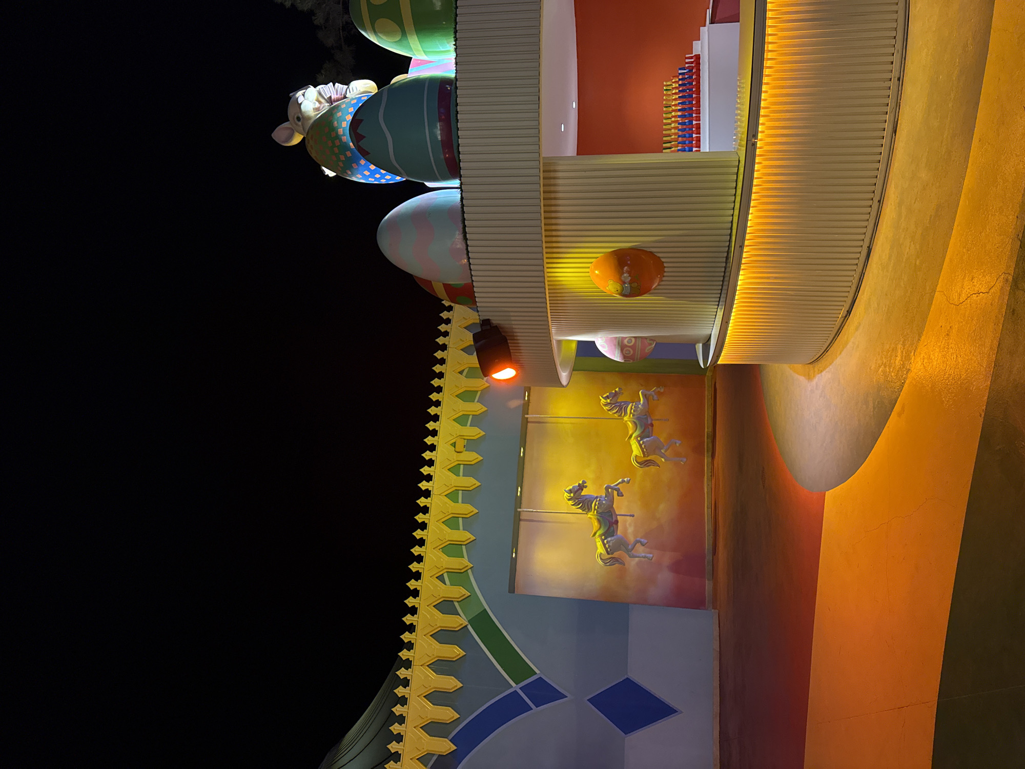The image depicts an outdoor area at night, likely an amusement park or entertainment venue, with a distinctly festive and whimsical theme. A central, curved counter structure with vertical white paneling is prominently featured, illuminated from beneath by warm yellow lights, casting a glow onto the textured ground. Above the counter, several large, vibrantly colored Easter egg decorations are visible, one topped by a large, stylized bunny figure dressed in a patterned blue and yellow garment. The bunny appears to be waving.
To the left of the counter, a mural adorns a wall, depicting two white carousel horses in mid-gallop against a bright yellow and orange background. The wall itself features a colorful, geometric design with a yellow, jagged border along the top and blue diamond shapes.
The ground in the foreground is a smooth, paved surface marked with distinct color blocks of dark green, orange, and a lighter yellow-orange, suggesting a pathway or designated area. The overall lighting is dramatic, with strong contrasts between illuminated areas and the dark night sky, hinting at an event or attraction that is actively lit for evening viewing. The context of Bodrum, Turkey, suggests this might be a seasonal attraction or part of a larger resort's entertainment offerings. No individuals are visible in the image, and no actions or interactions are taking place. The scene is static, focusing on the decorative elements and architectural features of the location.
