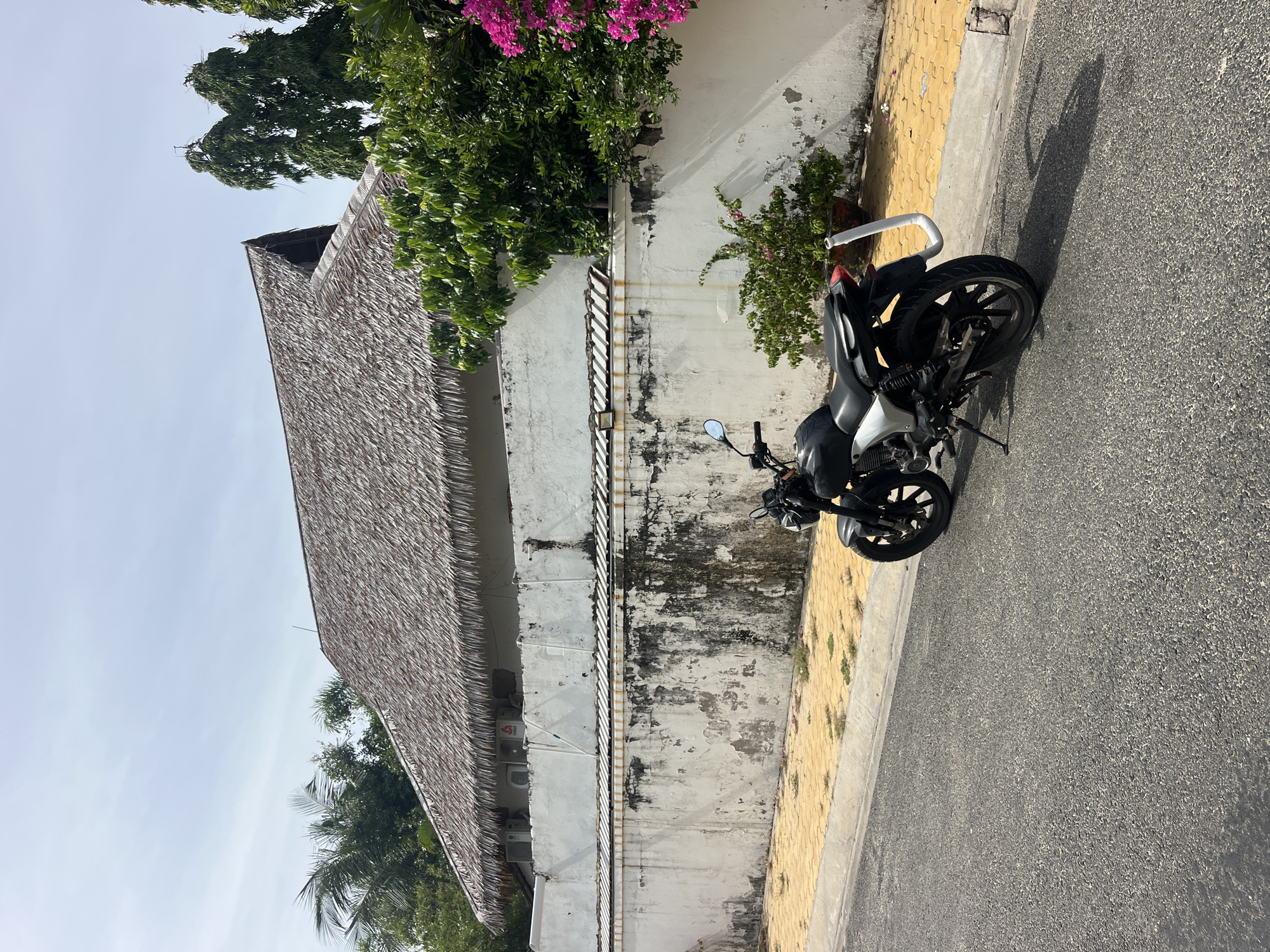 A black motorcycle is parked on the side of an asphalt road in Mũi Né, Vietnam. The motorcycle is positioned on the road, with its kickstand down. Behind the motorcycle, a white wall with weathered patches and dark stains runs along the property line. Above the wall, a building with a thatched roof is visible, and two air conditioning units are mounted on the wall below the roofline. Lush green trees and vibrant pink bougainvillea flowers grow near the building and wall. The ground next to the wall is paved with yellow tiles, and sparse patches of grass grow between the tiles. The sky is clear with some wispy clouds.