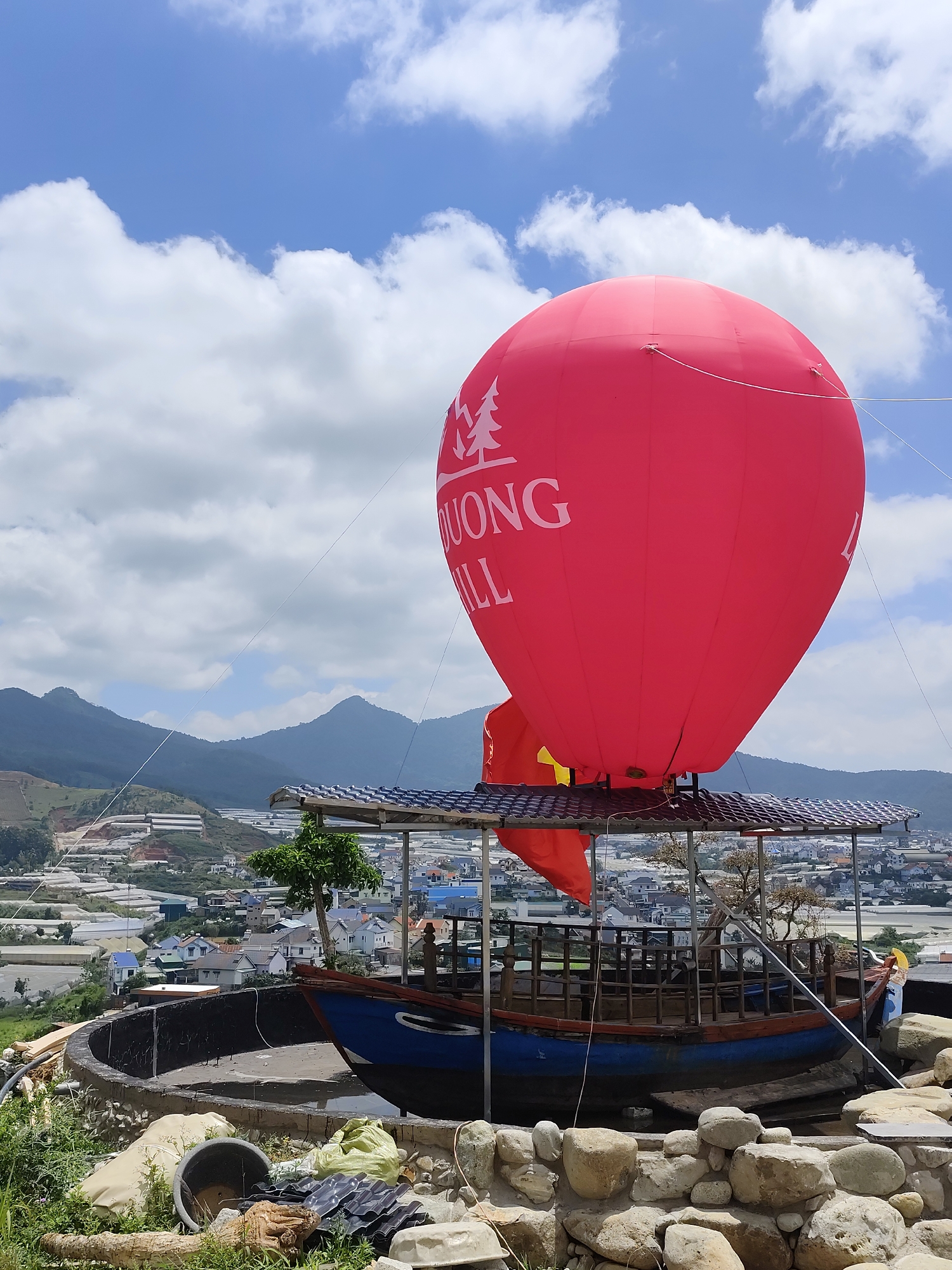 The image displays a large, vibrant red hot air balloon installation set against a clear blue sky with scattered white clouds, overlooking a valley in Lạc Dương, Vietnam. The balloon, which appears to be a stationary attraction rather than functional, features a white tree logo and the partially visible text "DUONG HILL."
Beneath the balloon, a rustic wooden boat with a blue hull and natural wood interior serves as its gondola, covered by a dark metal roof structure. The boat is positioned within a circular, concrete-lined basin. In the immediate foreground, a rough stone wall separates the viewer from the boat, with miscellaneous items like a black bucket, sacks, and corrugated metal sheets visible among green weeds. A small, sparsely leafed tree stands near the boat.
The background reveals a panoramic view of mountainous terrain. Distant mountains frame a valley filled with numerous white greenhouses or poly-tunnels, indicating agricultural activity typical of the region. Below these, a small town or settlement with various buildings is visible. The scene is brightly lit, suggesting mid-day sun, and conveys a sense of a scenic, perhaps tourist-oriented, location. There are no people visible in the image.