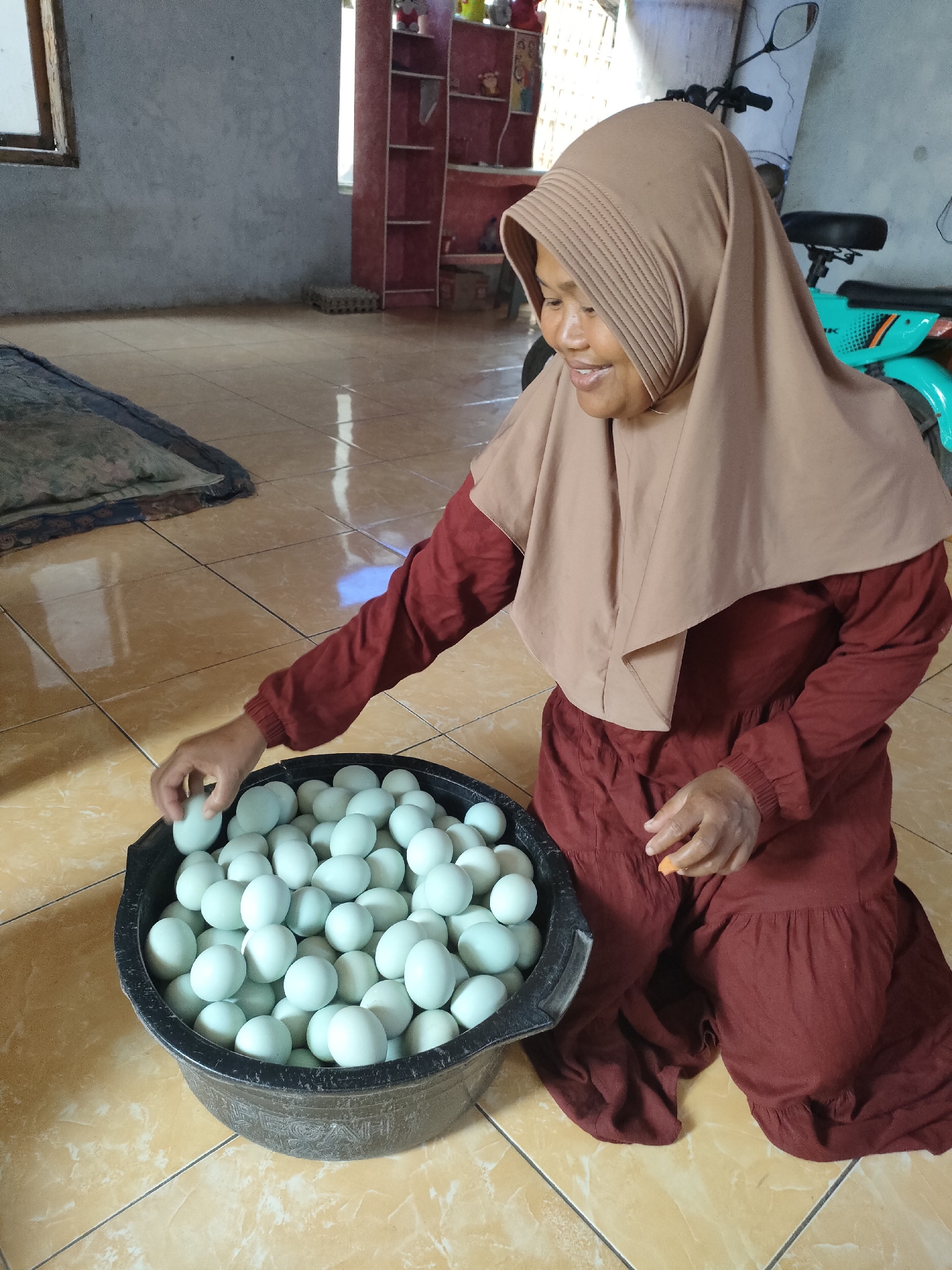 A woman, wearing a light brown hijab and a long maroon dress, kneels on a tiled floor in what appears to be a home or storage area in Tongaswetan, Indonesia. She is sorting through a large black basin filled with numerous light blue-green eggs. Her right hand is holding one egg, while her left hand is poised over the basin. In the background, a shelf unit with red accents is visible, along with a portion of a teal and black scooter. The environment is illuminated by natural light, casting reflections on the glossy floor tiles.