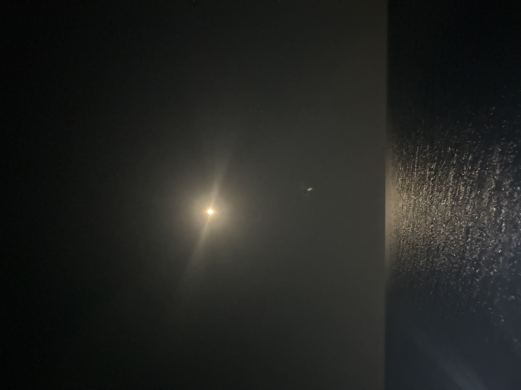 The image shows a moonlit sea at night. The moon is a bright orb in the dark sky, with a lens flare emanating from it. A reflection of the moon shimmers on the surface of the water, creating a path of light. The horizon line is clearly visible, separating the dark sky from the dark sea. The location is identified as the Agios Tychona Community in Cyprus.