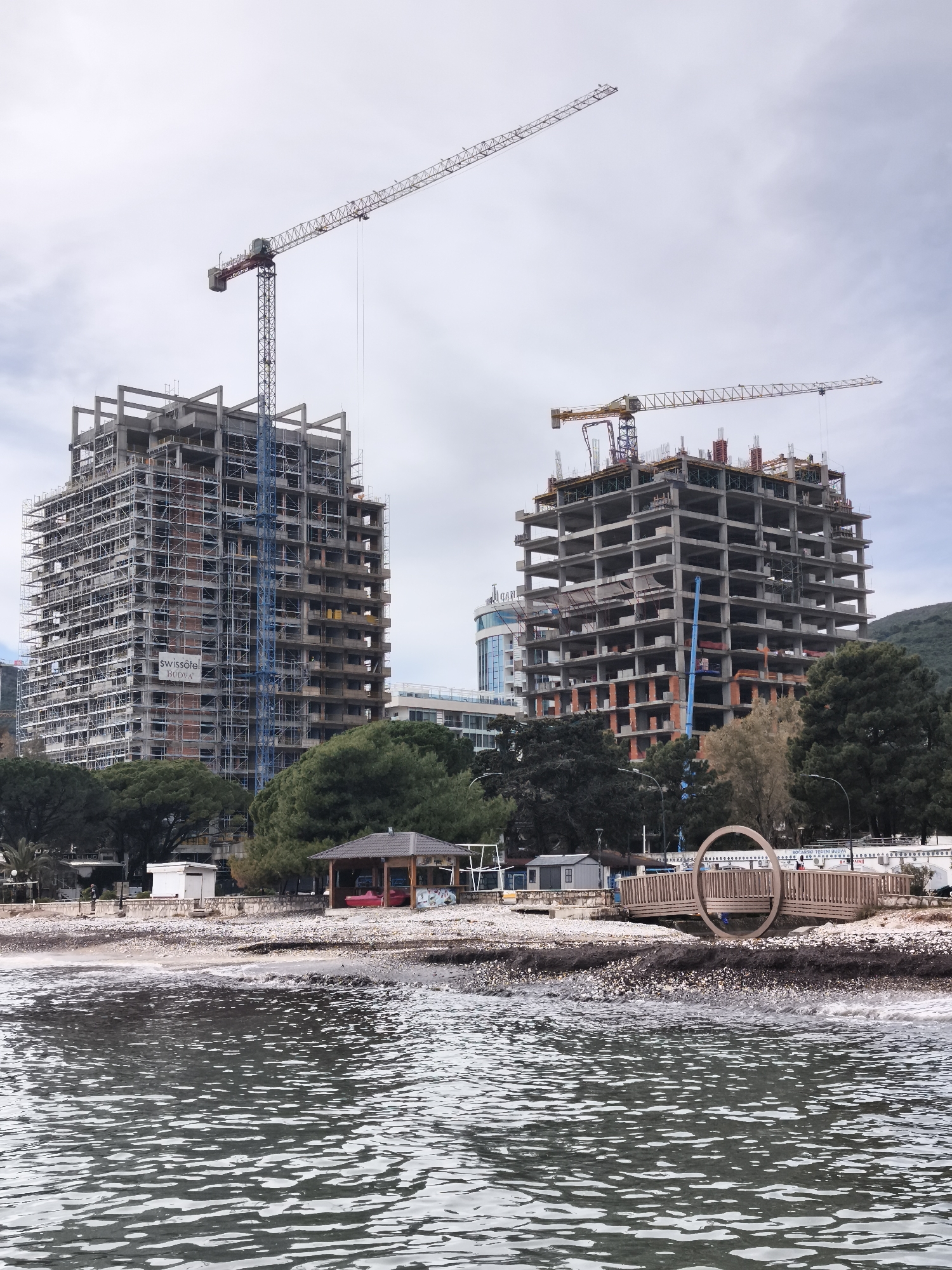 The image depicts a coastal urban scene in Budva, Montenegro, viewed from the water. The foreground consists of rippled, dark gray-green water transitioning to a pebbly beach littered with dark debris. Along the immediate shoreline, a small wooden walkway with railings forms a distinctive circular end feature. To its left, a small, light-colored rectangular shed stands, followed by a larger wooden gazebo with a dark roof, where red objects, possibly recreational equipment, are visible. A white wall with text including "BOCA.R.SKI TEČE.N.I BUDVA" is present further along the beach.
Behind the beach and a dense line of green trees, two multi-story concrete structures are under active construction. The building on the left is largely framed in concrete and extensively covered with scaffolding, displaying a "swissôtel BUDVA" banner on its facade. A tall red and blue tower crane is positioned next to this structure. The building on the right, also a multi-story concrete frame, appears less complete, with exposed slabs and columns, and is topped by a yellow tower crane. In the further background, a modern building with a curved glass facade is partially visible, and green, tree-covered hills rise. The sky is overcast with light gray and white clouds. No specific individuals or discernible actions are present.