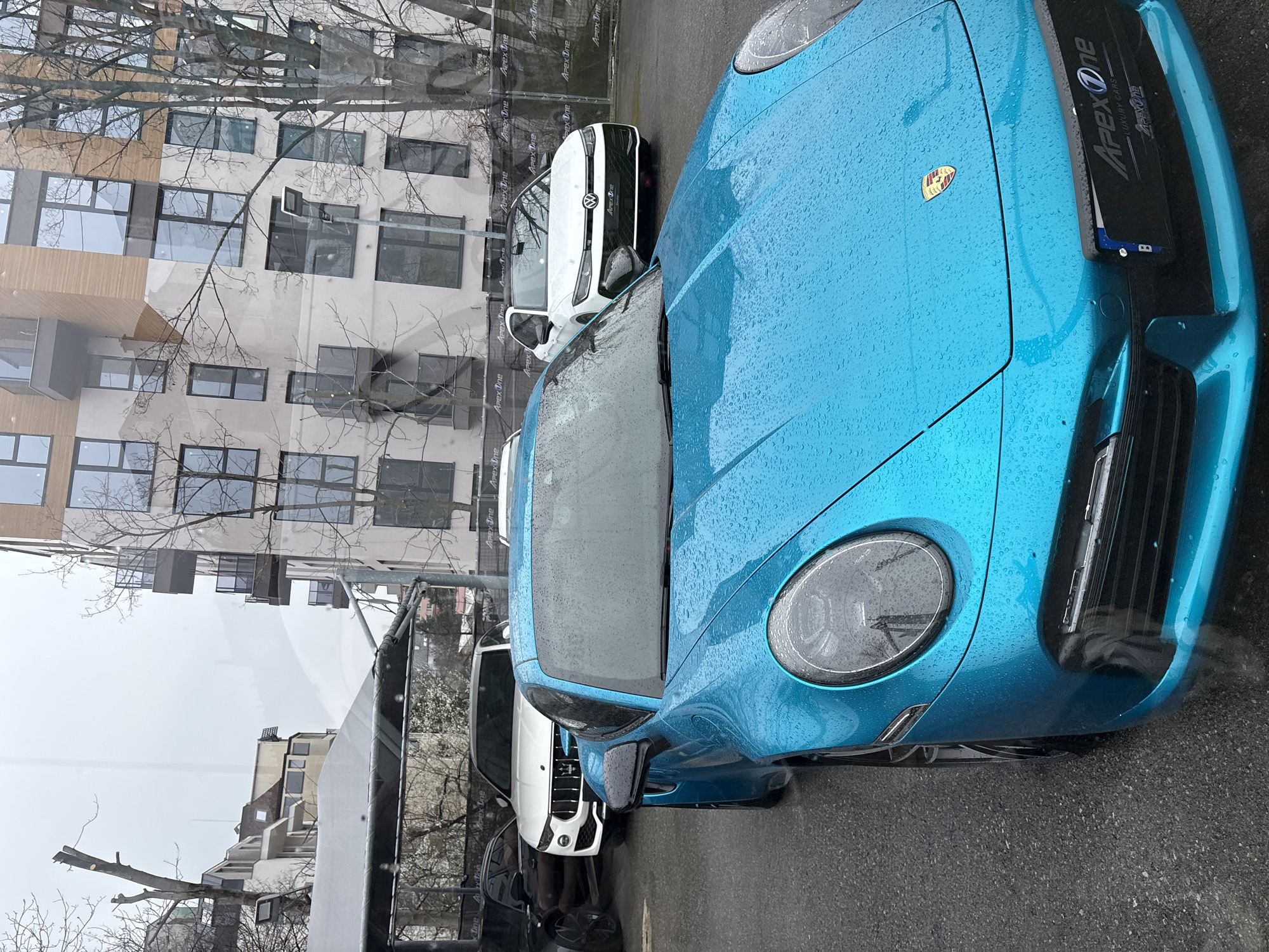 The image depicts a street scene in an urban environment. The primary subject is a bright blue Porsche sports car parked on the street, covered in raindrops. A white Volkswagen ID.3 is parked next to the Porsche. Behind the cars is a building with multiple windows and an "Apex One" sign. The photo appears to have been taken from a slightly elevated position, possibly from inside a building, looking out through a window. Based on the 'Apex One' sign and general architecture, the location is likely in the city of Sofia, Bulgaria. The buildings reflected on the Porsche show more buildings. The day appears overcast, indicated by the muted lighting and droplets on the car.