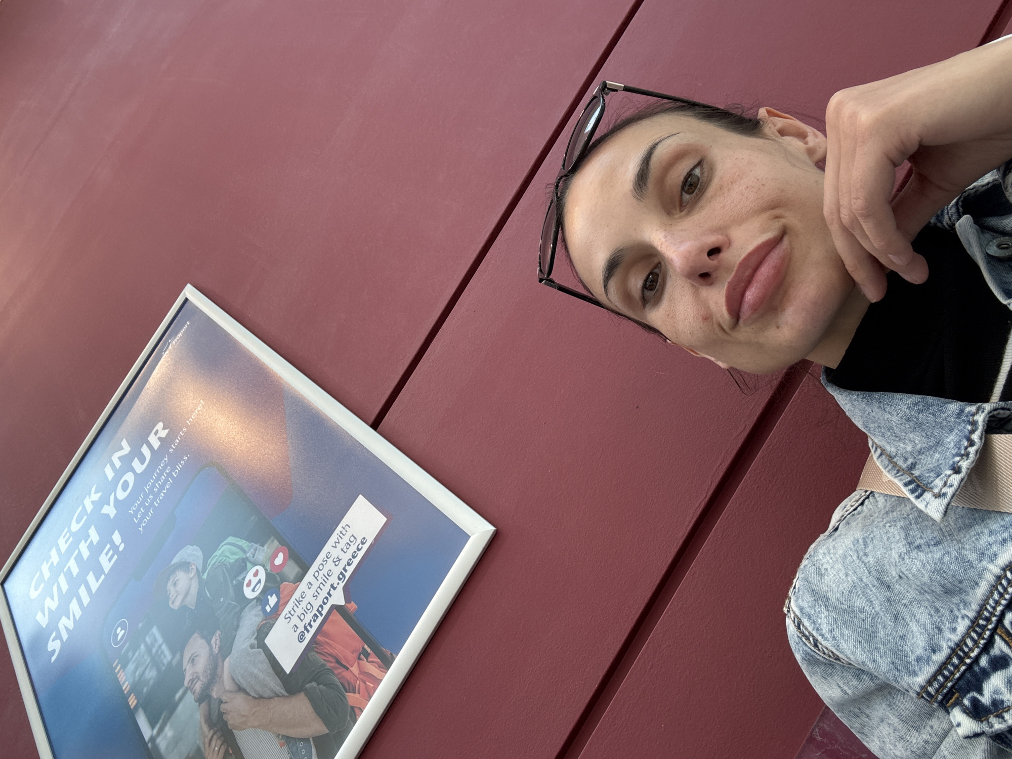 The image is a selfie taken by a woman wearing glasses on her head, a black top, and a light blue denim jacket with a tan strap across her shoulder. She is posing with her hand near her face, looking into the camera. To the left of the woman is a framed advertisement for Fraport Greece, encouraging travelers to "CHECK IN WITH YOUR SMILE!" and tag @fraport.greece. The background consists of a maroon-colored wall with vertical panels. There is no direct visual information to confirm the location is Mouzouras, Greece.
