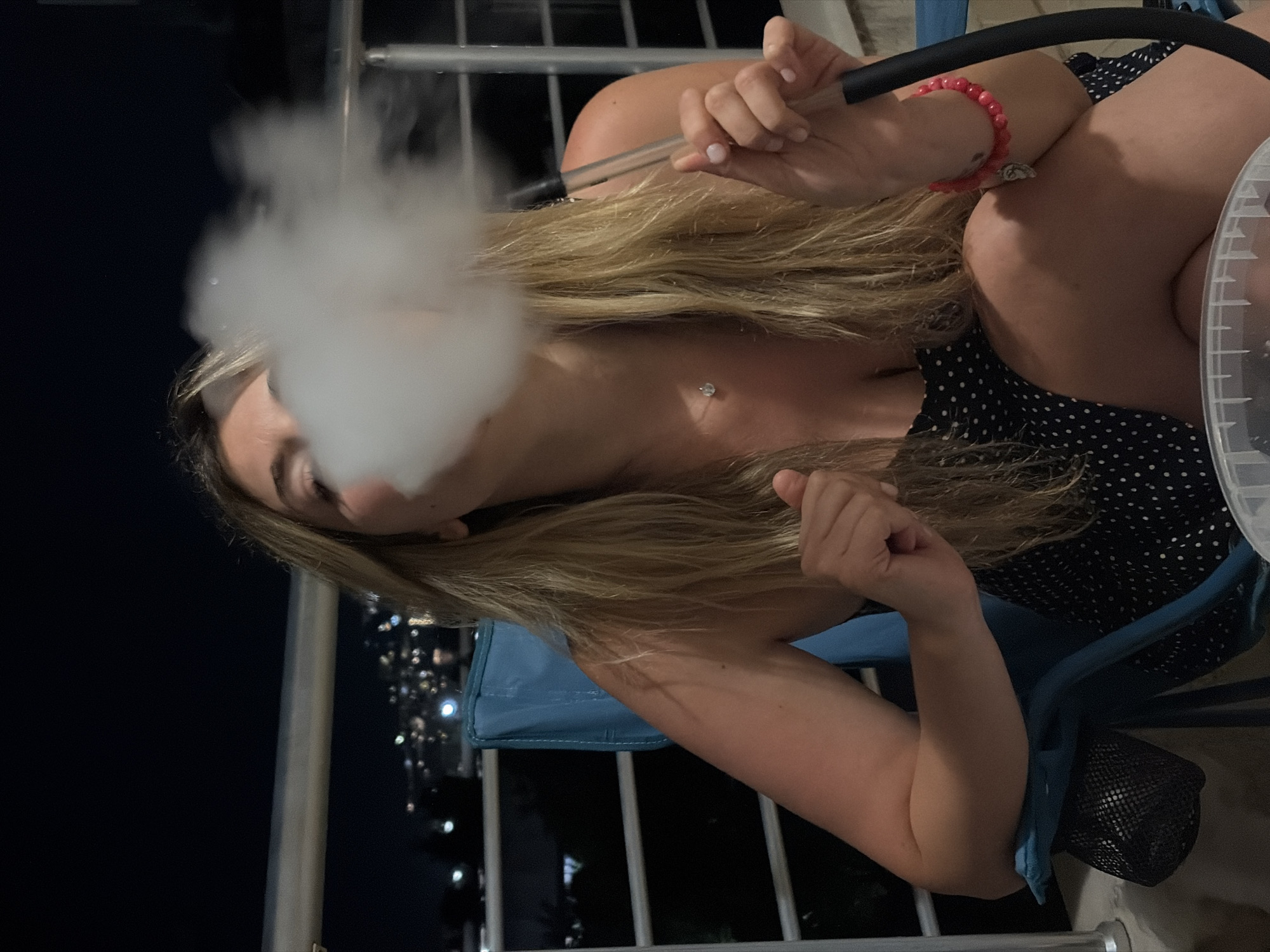 A woman with long blonde hair sits in a blue folding chair on a balcony with a railing, smoking a hookah. She wears a black and white polka-dot top and a red bracelet. Smoke obscures part of her face. The city lights of Budva, Montenegro, are visible in the background.
