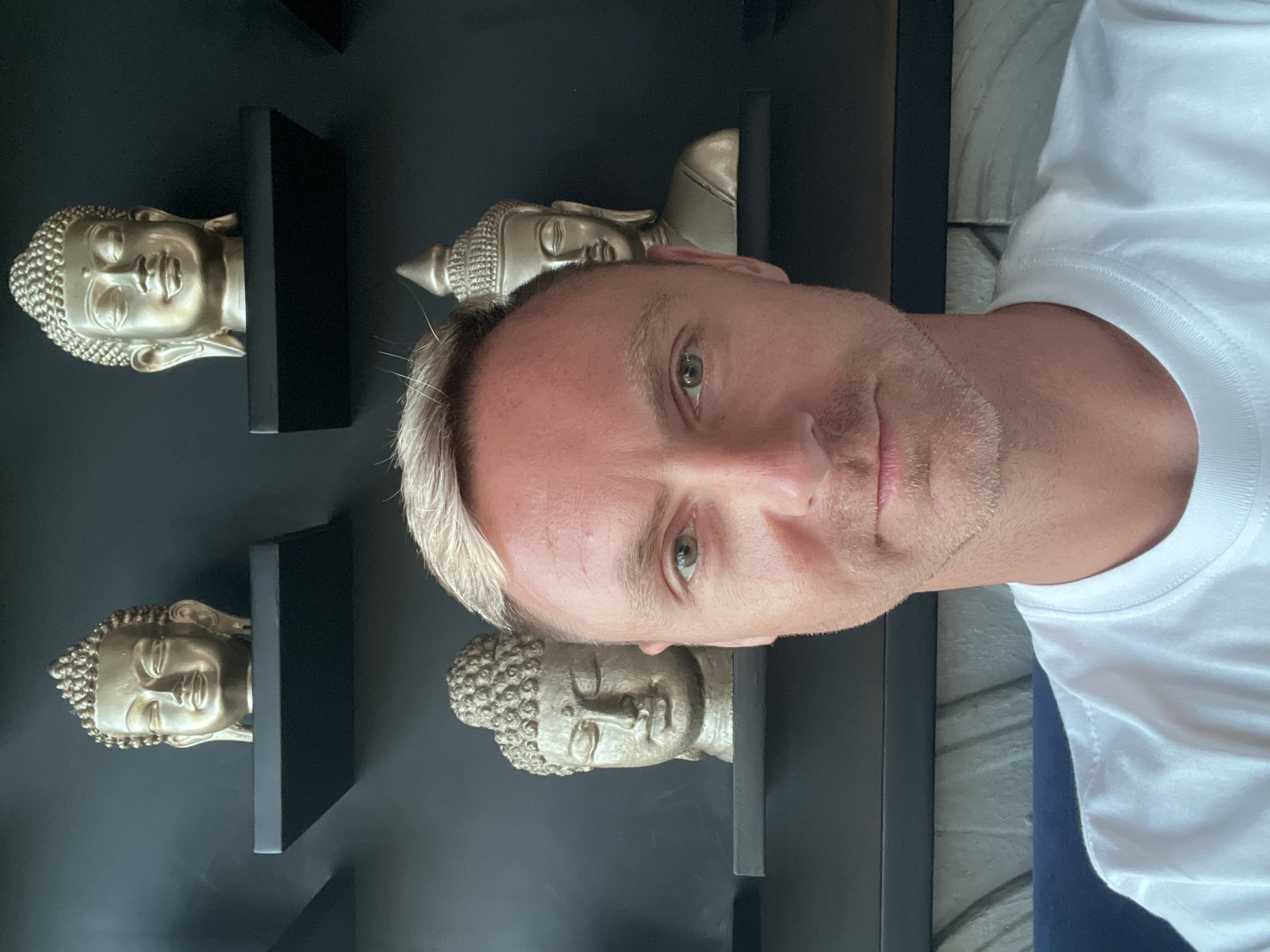 The image is a selfie of a man with blond hair and a white t-shirt, taken in a room with black shelves on a dark wall. There are several Buddha head statues on the shelves. The man is looking at the camera. The environment suggests an indoor setting, possibly a home or a hotel room, in North Kuta, Indonesia.