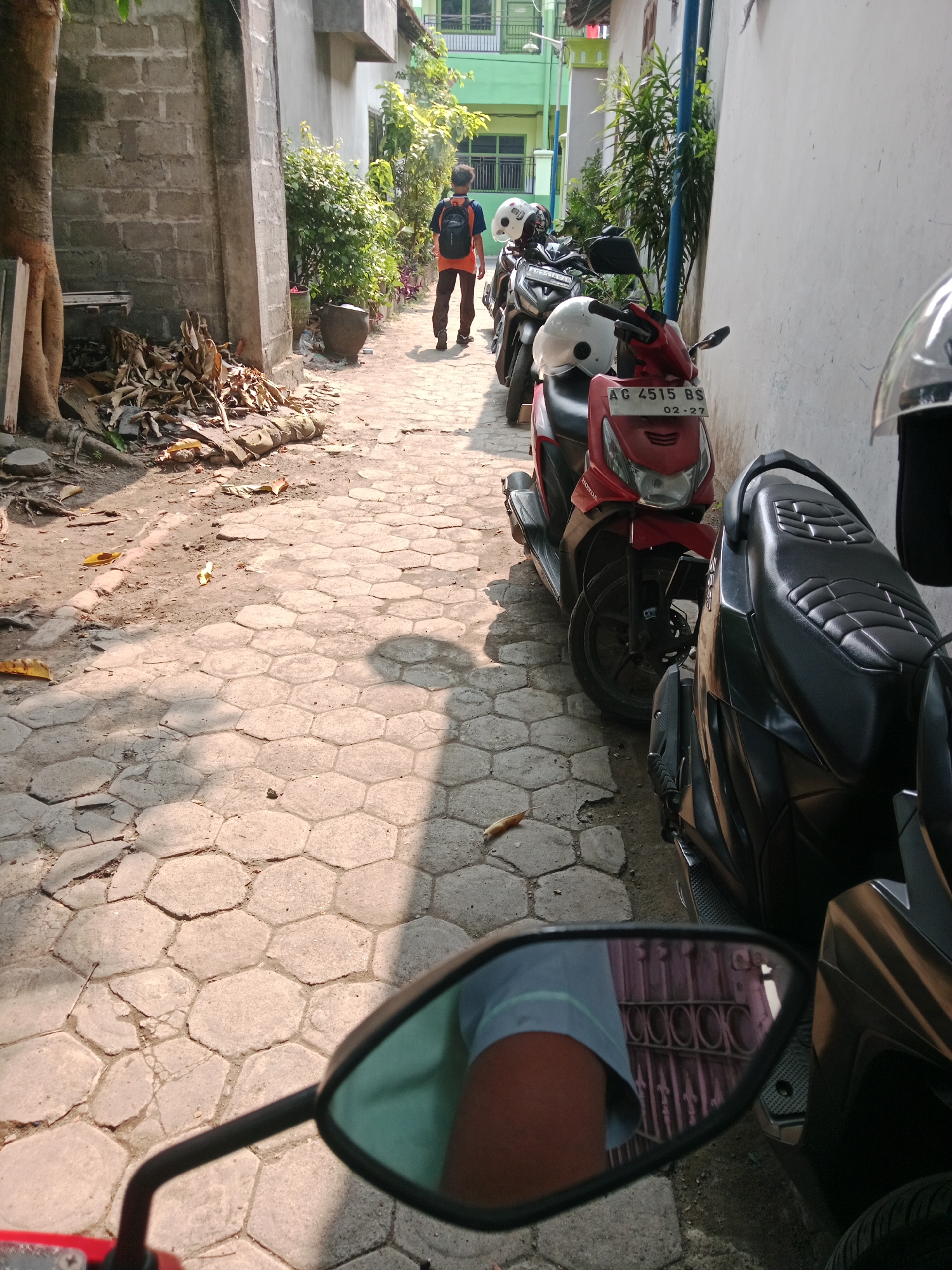 The image captures a narrow alleyway in the city of Kediri, Indonesia, likely a residential area. The ground is paved with hexagonal concrete tiles, and sunlight casts distinct shadows. To the left, a rough concrete wall borders the alley, with a large tree trunk and a pile of dry leaves at its base. A young person wearing a backpack and an orange shirt walks away from the camera. A row of parked motorcycles, primarily scooters, lines the right side of the alley, some with helmets placed on them. The motorcycles are parked closely together against a white wall. In the foreground, a motorcycle's side mirror reflects a partial view of the alleyway and a person's arm wearing a blue shirt. The scene suggests a typical daily scene in a neighborhood with limited parking space.