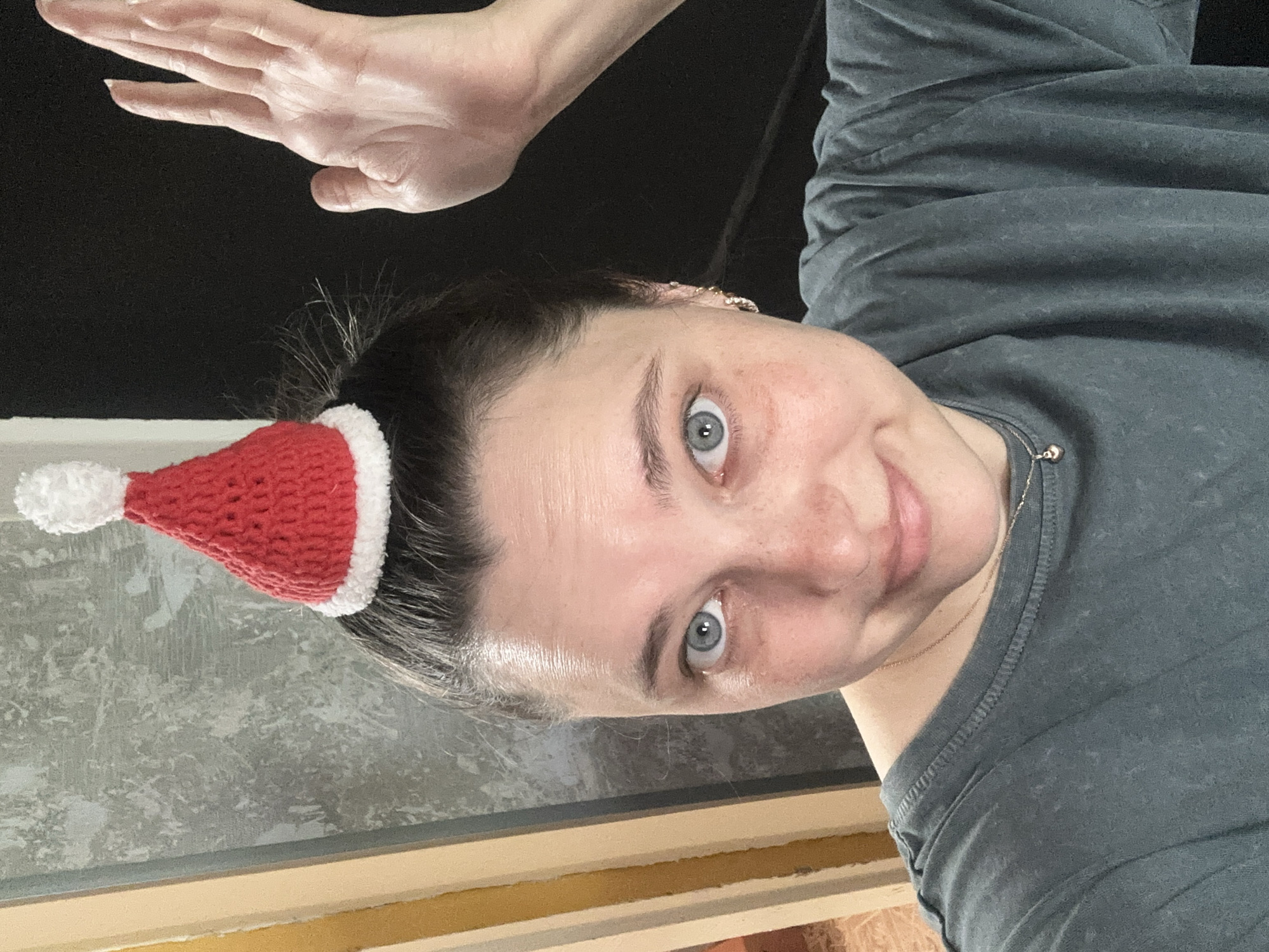 A woman with blue eyes and dark hair pulled back is wearing a small, red crocheted Santa hat with a white pom-pom and brim. She is looking at the camera and smiling, with her right hand raised in a wave. She is wearing a dark gray t-shirt and a delicate necklace with a small heart pendant. The background is blurred and appears to be an indoor setting with a textured wall and a black surface. There are no discernible landmarks or contextual elements to place this image in Yaroslavka, Russia, or any other specific location. The scene is a close-up portrait of the woman.