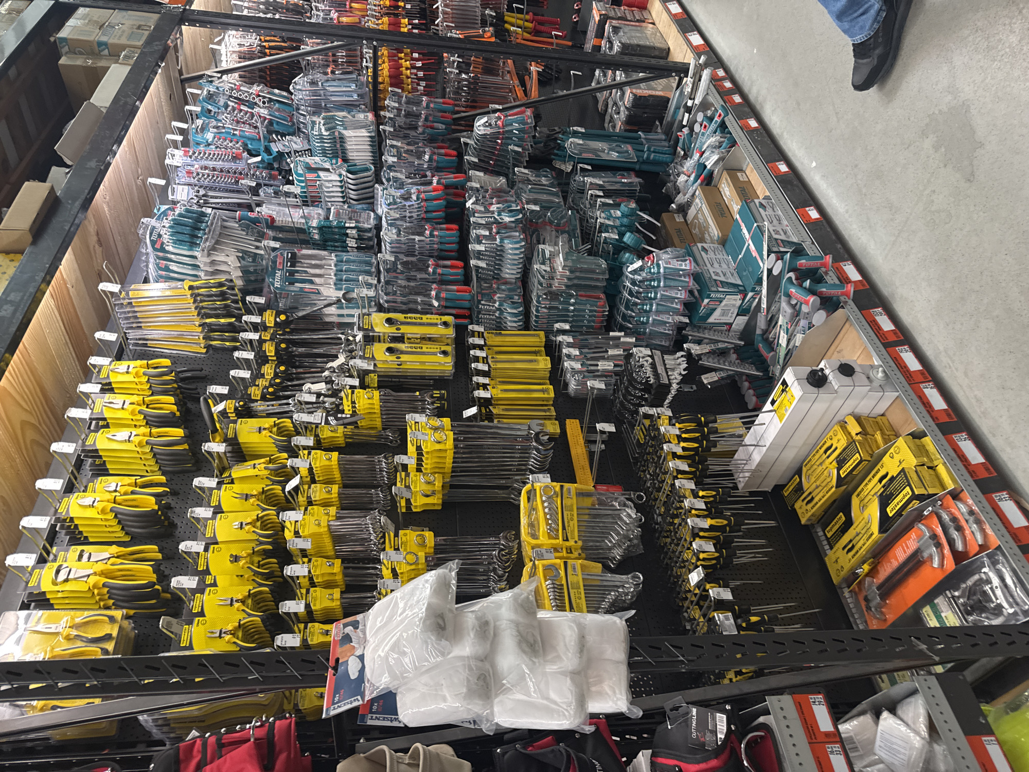 This is an overhead shot of a hardware store shelf stocked with an array of tools. The tools are neatly arranged and grouped by type, featuring yellow-handled pliers, wrenches, screwdrivers, and tool sets. There are also blue-handled tools. The shelves are black metal and appear to be well-organized. In the bottom of the frame, there are plastic bags, a red bag and the corner of a gray bag. In the upper right corner, a shoe is visible on a gray floor. Some of the packaging on the tools indicates the brand "Total" and "Topmaster". Based on the OCR, there is also a "WISENT" label. The setting is presumably in Sofia, Bulgaria.