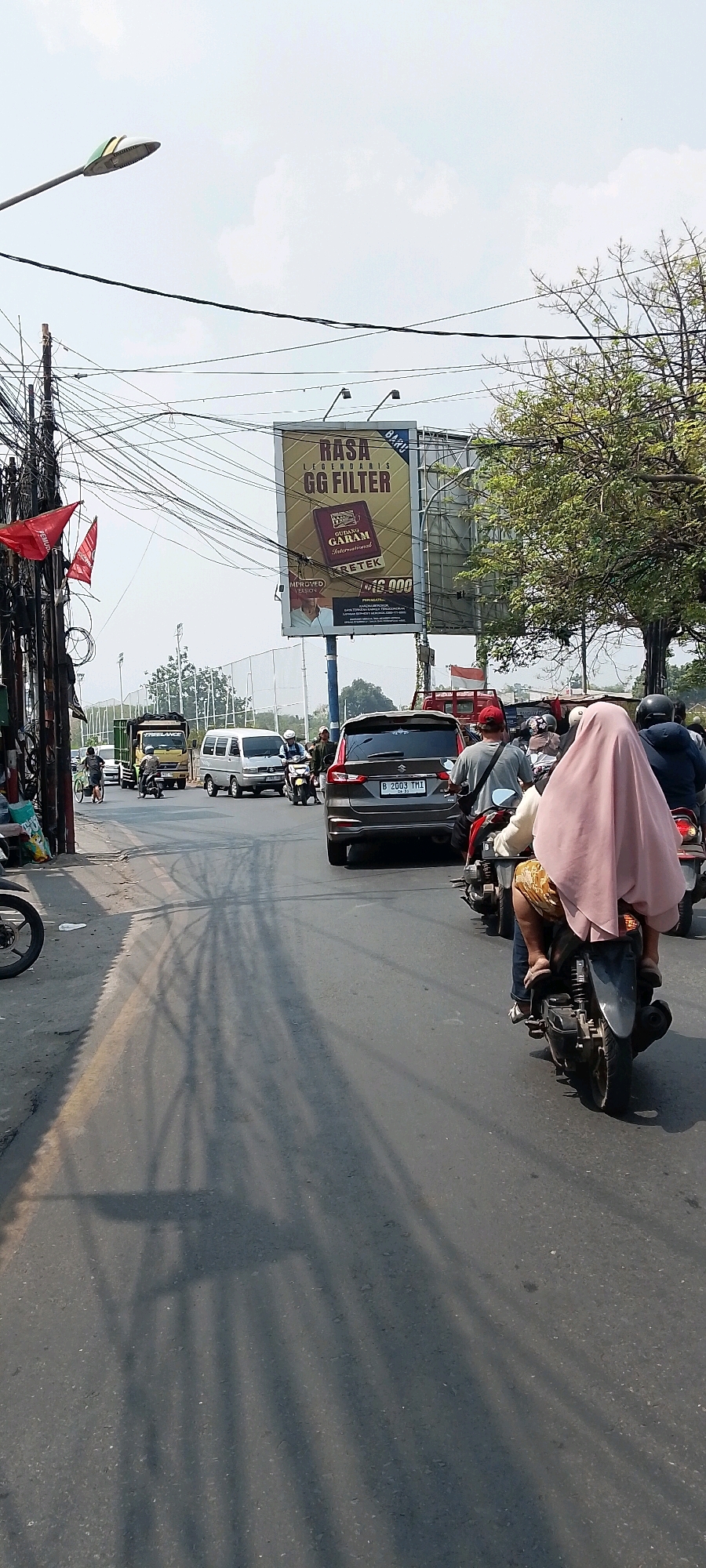 This image captures a street scene in Bekasi, Indonesia, likely on a sunny day with clear skies. The perspective is from a moving vehicle, looking forward and slightly to the right. The foreground is dominated by a road surface showing tire marks and shadows from overhead wires and possibly trees.
In the middle ground, several vehicles are visible on the road. A dark-colored SUV with license plate B 2003 TMI is in the right lane. Ahead of it, a white van and a yellow truck are in the left lane. Several motorcycles are also present, with riders, including one prominently featuring a person wearing a pink headscarf on the back of a motorcycle in the foreground.
On the left side of the road, there's a dense collection of utility poles and tangled electrical wires, a common sight in urban areas. Two red flags are attached to a pole, bearing some text that is partially obscured. To the right of the road, a large billboard is visible, advertising "RASA LEGENDARIS GG FILTER" and "GUDANG GARAM INTERNATIONAL CRETEK," with a price of Rp 16.000. Beyond the billboard and the vehicles, a sports field with fencing and trees can be seen. The overall impression is of a busy, everyday street in an Indonesian city.
