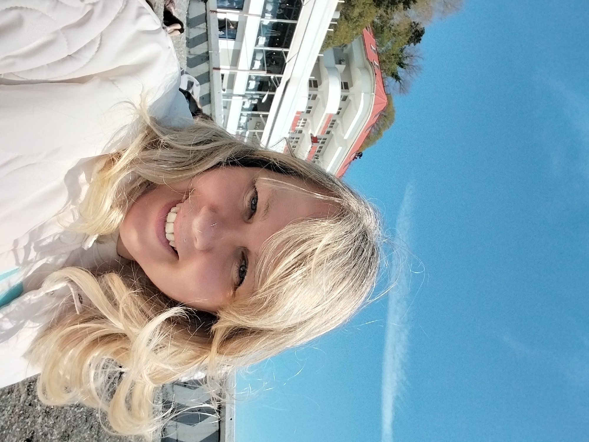 A close-up, rotated portrait captures a smiling woman with long, wavy blonde hair, looking directly at the camera. The image is oriented sideways, with the woman's head tilted towards the bottom left. She wears a white quilted jacket, with a glimpse of light blue fabric visible near her collar. A small, subtle piercing is visible on her nose. Her smile is broad, revealing her teeth, and her blue eyes appear happy.
The background on the left side of the frame features a bright blue sky, clear except for a faint, wispy white cloud or contrail. On the right, behind her, stands a large, multi-story building with white walls, numerous windows, and a distinctive red-tiled roof. The architecture suggests a modern resort or hotel, partially obscured by some green trees and foliage. The bright natural lighting indicates a sunny day. This outdoor scene is set in Sochi, Russia. No visible text is present in the image.