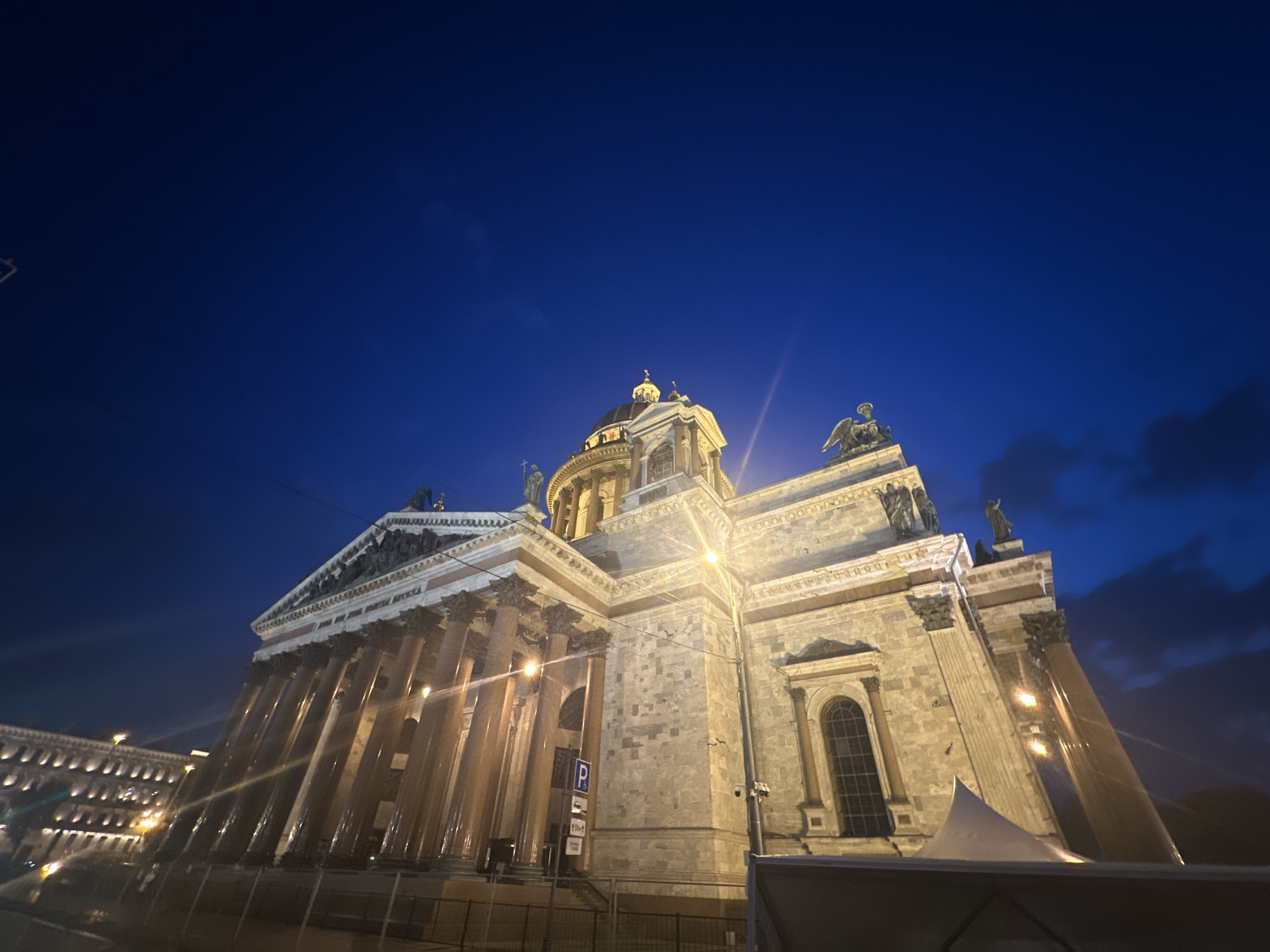 The image shows Saint Isaac's Cathedral in Saint Petersburg, Russia at night. The cathedral is illuminated with warm, golden light, contrasting with the deep blue of the evening sky. The structure is viewed from a low angle, emphasizing its size and grandeur. Details visible include the cathedral's dome, columns, statues on the roofline, and a parking sign. The scene seems to be a public space near the building.