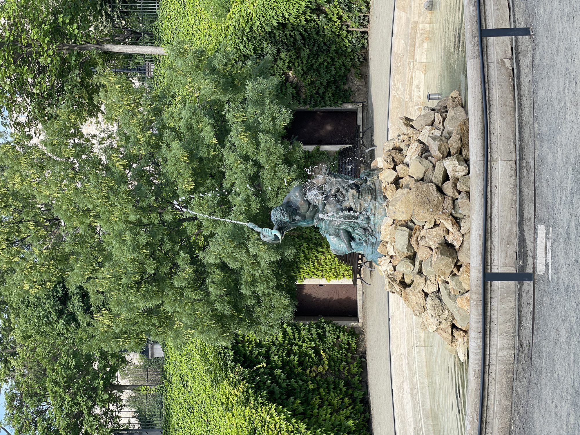 This image shows the Matthias Fountain in Budapest, Hungary. The fountain features a bronze sculpture of King Matthias Corvinus hunting. The sculpture depicts the king on horseback, holding a crossbow. The fountain is surrounded by rocks and lush greenery, with a building covered in ivy in the background. A wrought iron fence is visible in the distance, partially obscured by trees. The setting is an outdoor urban environment, likely a park or public square. A street runs alongside the fountain.