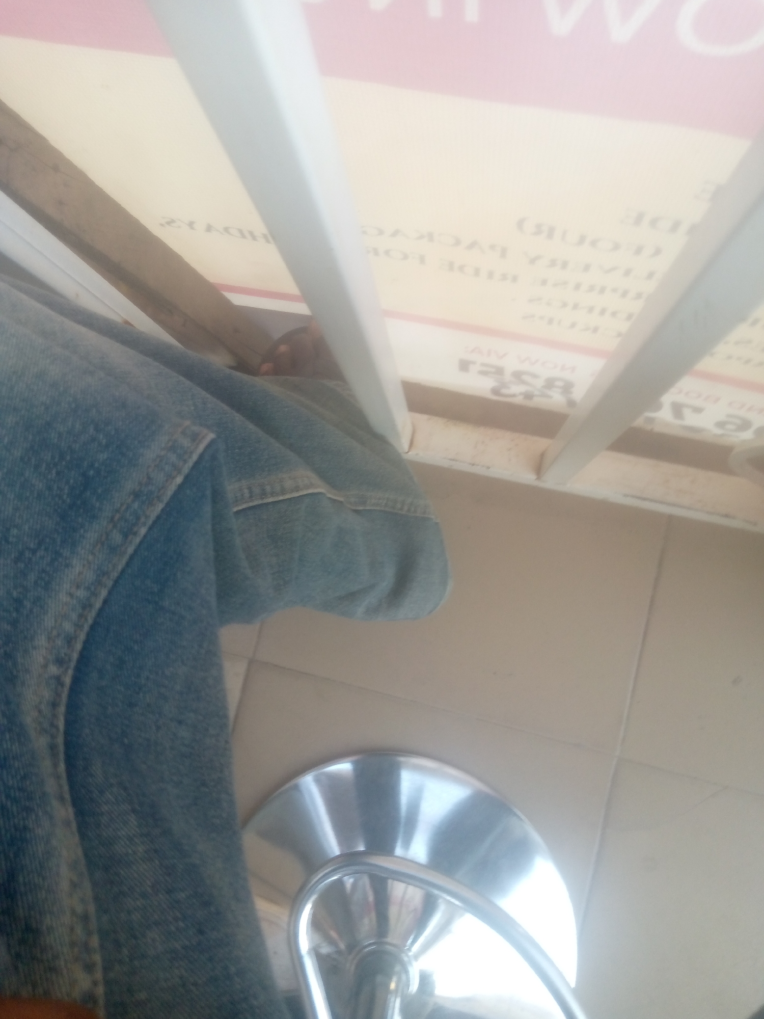 A person, partially visible, is sitting with legs crossed, wearing blue denim jeans. They are positioned under a light-colored surface with text, suggesting a commercial establishment or public area. The surface is supported by white vertical beams and a wooden beam. In the foreground, a portion of a silver-colored stool or chair base is visible, reflecting light. The background consists of light-colored tiled flooring. The scene appears to be indoors, possibly in a shop or waiting area in Jalingo, Nigeria. The text on the surface is largely obscured and appears to be in English, with some words or phrases readable, such as "RIDE", "PACKAGES", and potentially a business name.
