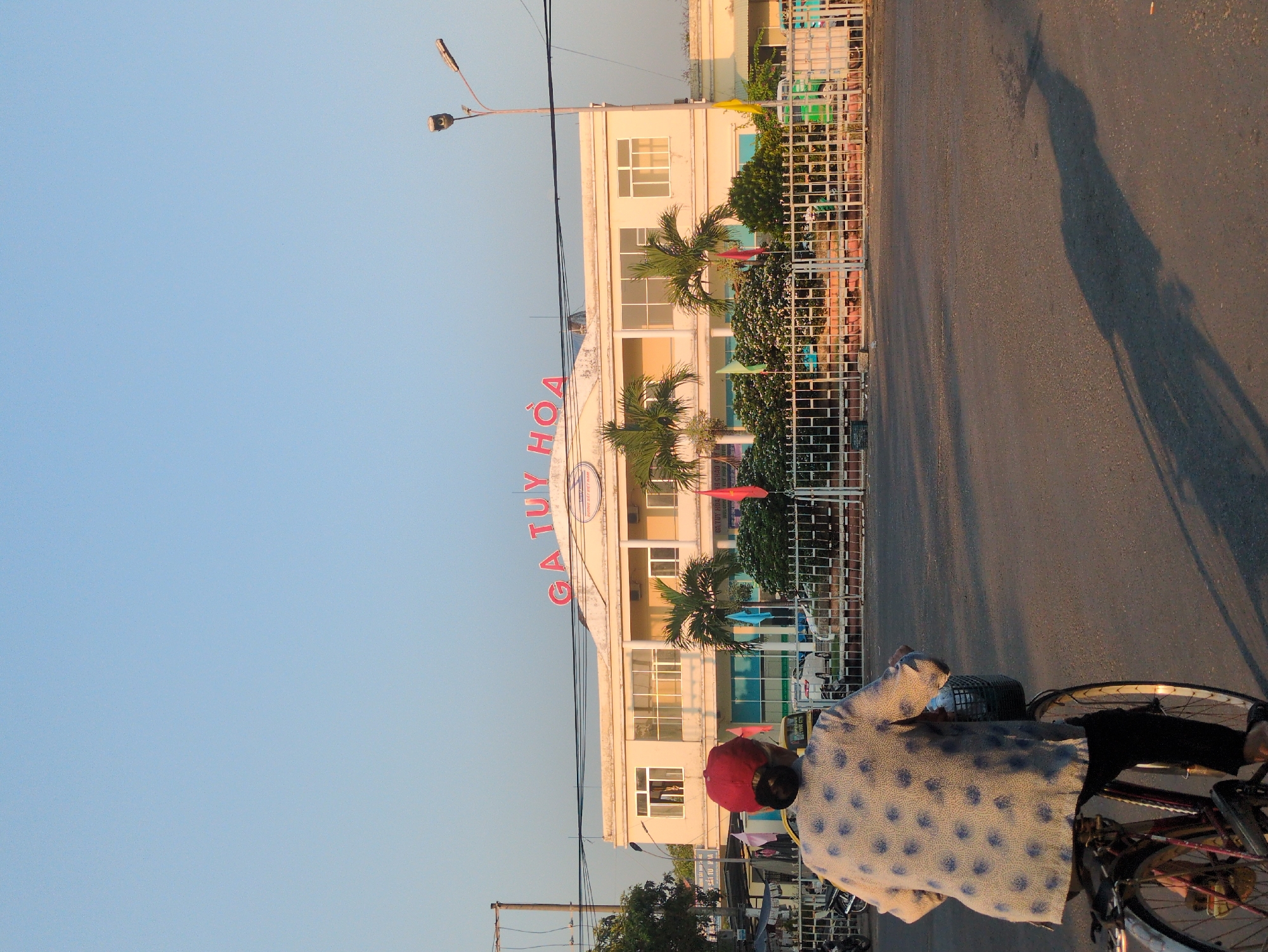 The image captures a scene in Tuy Hòa, Vietnam, featuring a person riding a bicycle on an asphalt road. The rider is wearing a light-colored shirt with a pattern of dark blue polka dots and a red cap. The bicycle is a traditional model.
Alongside the road is a large, multi-story building with the words "GA TUY HOA" in prominent red lettering on its facade. The building is partially obscured by a fence. Palm trees and other greenery are visible in front of the building, adding to the tropical atmosphere. Utility poles and wires run overhead. The sky is a clear, gradient blue. The sun is low in the sky, casting a long, dramatic shadow of the cyclist and their bicycle onto the road.