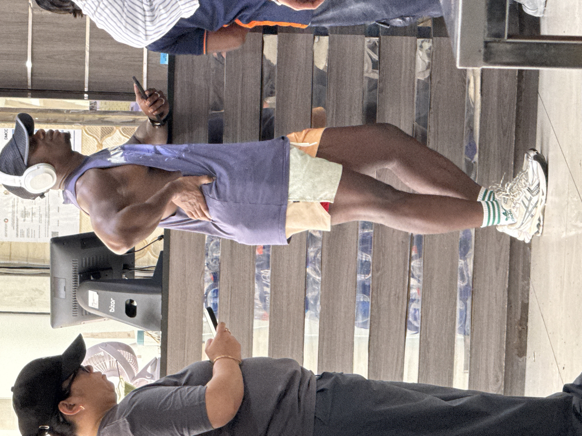 The image depicts a scene with at least three individuals in what appears to be a public indoor setting, possibly a reception area or waiting room. In the foreground on the left, a person wearing a dark baseball cap and a grey t-shirt is holding a smartphone. To the right of this person, a man with white headphones on and wearing a sleeveless purple tank top and multicolored shorts is standing and looking to his right, with his right hand on his hip. He is also holding a smartphone in his left hand. Behind him, another person is partially visible, wearing a blue shirt with orange trim. In the background, a computer monitor on a stand with the logo "Bizor" is visible, along with other documents and what appear to be shelves with bottles of water. The environment has a tiered wooden counter or structure. The flooring is tiled. The overall impression is that of a casual encounter or waiting period in a commercial or service-oriented location within Dubai, United Arab Emirates.