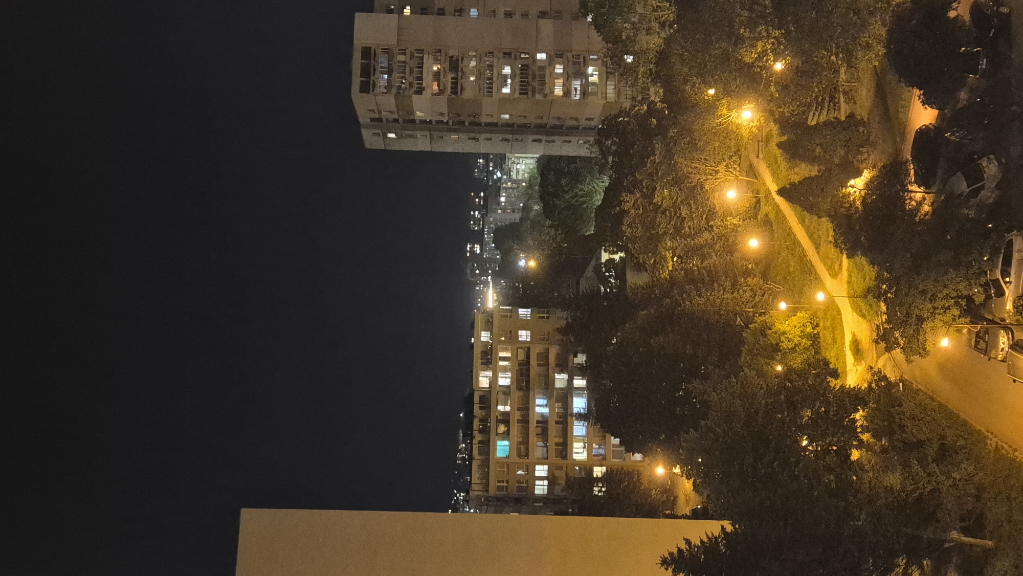 This media file captures a nighttime view of a residential area in Varna, Bulgaria, taken from an elevated perspective. The scene, which appears to be rotated 90 degrees clockwise, depicts an urban landscape after dark.
In the foreground, a light-colored building or wall is partially visible at the bottom. To the right, a path or narrow road winds through an area dense with trees and bushes. This path and the foliage are brightly illuminated by a series of warm yellow streetlights, casting distinct shadows and highlights. Several vehicles, including what appears to be a van, are partially visible parked or driving along the illuminated areas.
The midground and background are dominated by large, multi-story apartment buildings. These structures feature numerous windows, many of which are lit, indicating occupancy and casting a warm glow. A few windows exhibit a cooler, blueish light. Further in the distance, more buildings with scattered lights are visible against the dark night sky, suggesting a sprawling urban environment.
The time of day is clearly night, evidenced by the completely dark sky and the reliance on artificial lighting from streetlights and building interiors. There are no people visible, and no specific activities or events are taking place; the scene is static and quiet. No text is visible in the image. The overall impression is that of a calm, illuminated urban residential area after dark.
