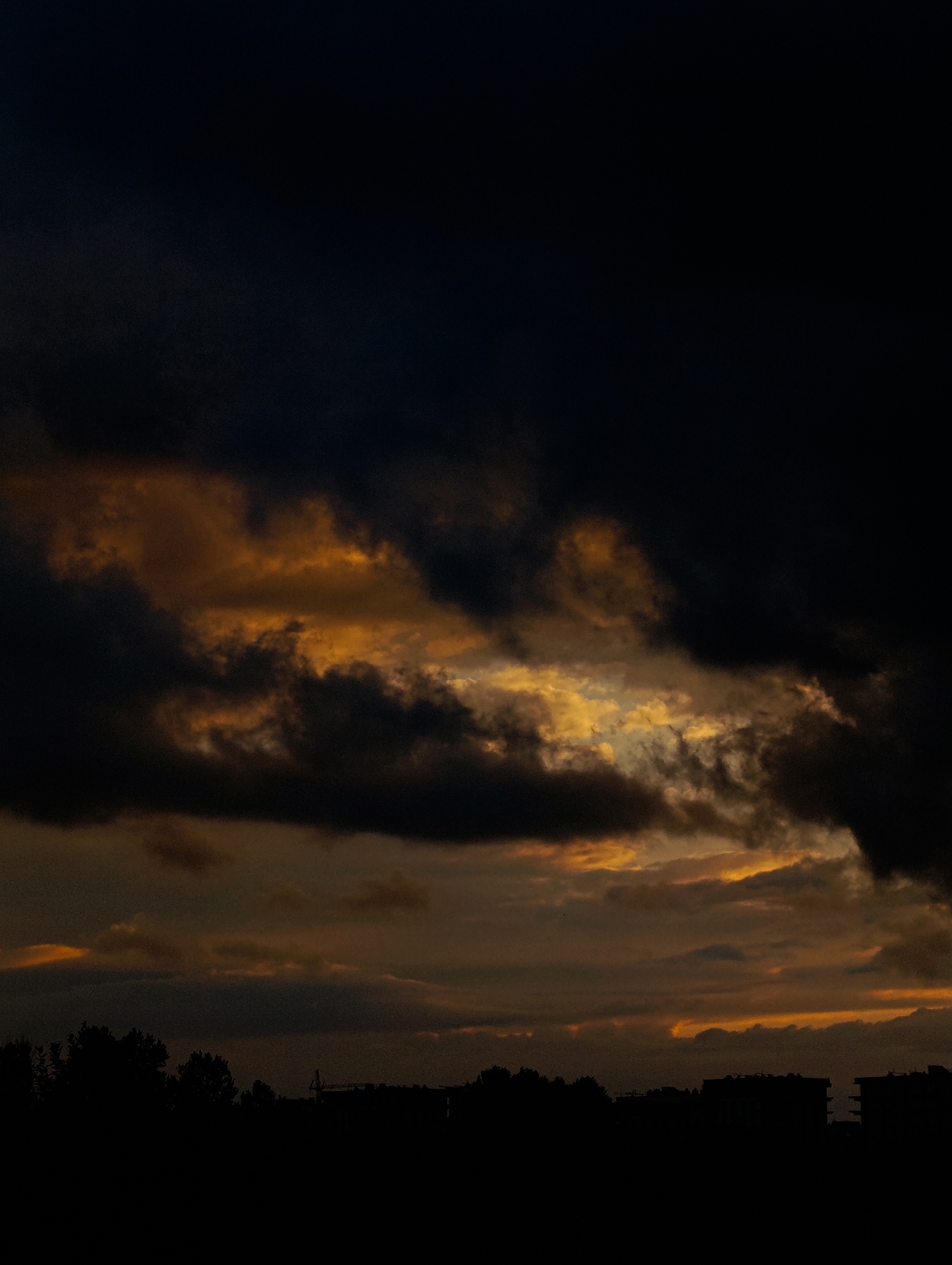 The image depicts a dark, brooding sky filled with dramatic clouds, likely during either sunrise or sunset. The clouds are a mix of deep blacks and muted oranges and yellows, suggesting the sun's light is filtering through them from a low angle. Below the sky, a silhouetted line of trees and buildings forms the horizon. In the distance, the faint outline of a construction crane is visible, hinting at development or ongoing projects within the city. The overall mood of the image is somber and atmospheric.