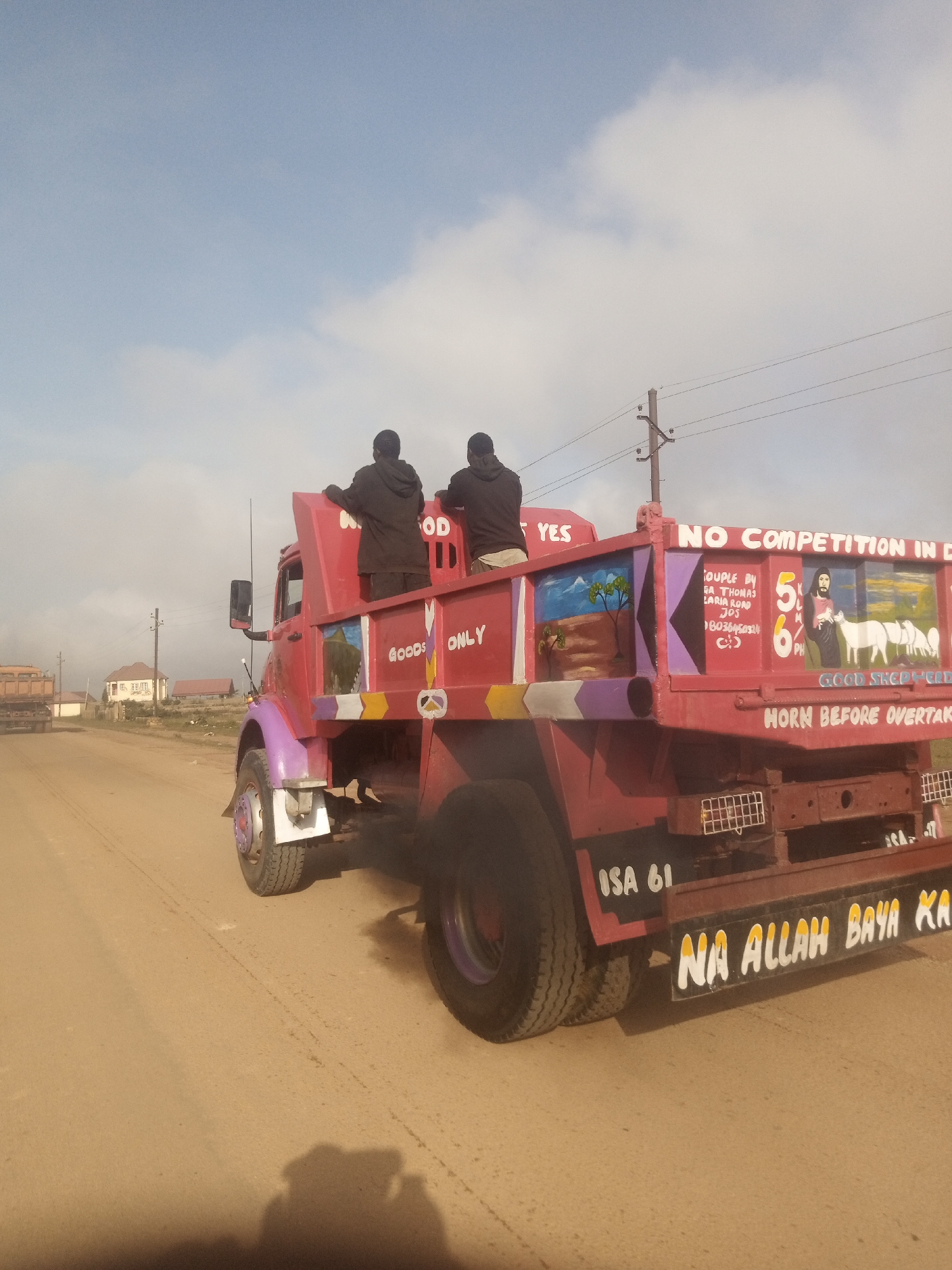 A medium shot captures the side of a red truck in Kwang, Nigeria, with two individuals standing in the back, facing away from the camera. The truck is decorated with paintings, including religious imagery and text in English and what appears to be a local language. The phrase "NO COMPETITION IN D" is visible on the upper rear of the truck, along with a depiction of Jesus with sheep. Below, "HORN BEFORE OVERTAKE" is painted. The truck's license plate is partially obscured but shows "ISA 61". The truck is on a dirt road, with a second truck in the distance on the left. The environment appears to be semi-arid, with a few buildings and power lines visible in the background under a pale blue sky with clouds. The lighting suggests it is daytime.