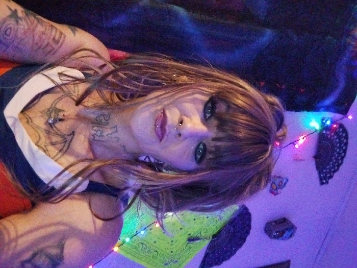 A woman with shoulder-length brown hair and bangs, laying down with her head tilted, poses for the camera. She has a nose ring and visible tattoos on her chest, neck, and arms. She wears a dark top with white trim. The background features colorful lights, decorative fans, a green cloth, and other objects. The overall lighting in the photo gives a blue hue.