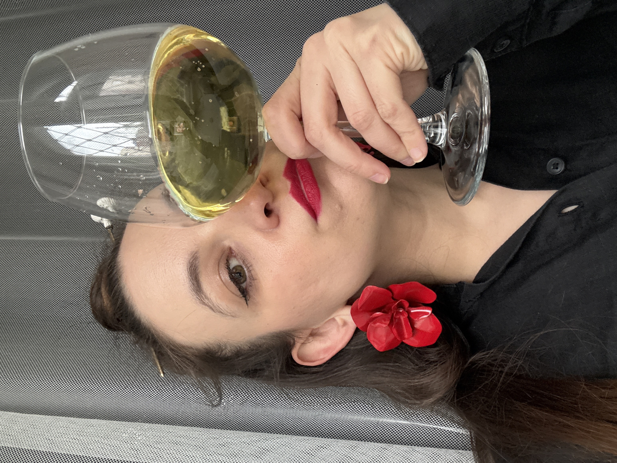 A woman with dark hair and red lipstick is lying on her side with a glass of amber liquid positioned over her lips. She is wearing a black shirt and has a red flower clip in her hair near her ear. The background appears to be a gray, mesh-like surface. There is no information about the location being Chaikovsky, Russia in the image.