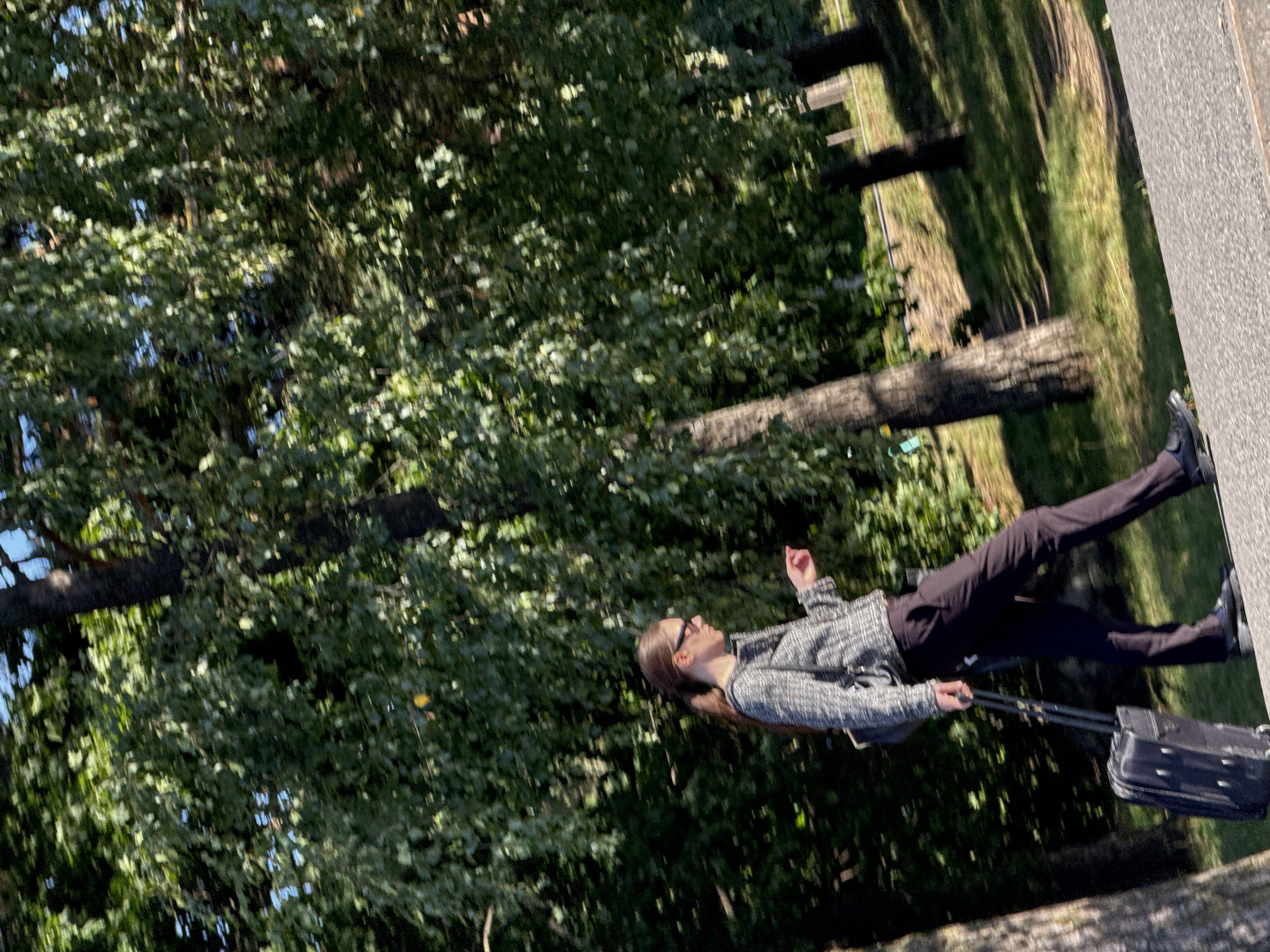 A woman walks with a rolling suitcase on a paved path next to a grassy area with trees. The woman is wearing a grey and white patterned jacket, dark pants, and black shoes. She is also wearing sunglasses and has her hair pulled back in a ponytail. The trees are green and leafy, and the sun is shining. The path appears to be in a park-like setting. The location is the Kadriorg Park in Tallinn, Estonia.