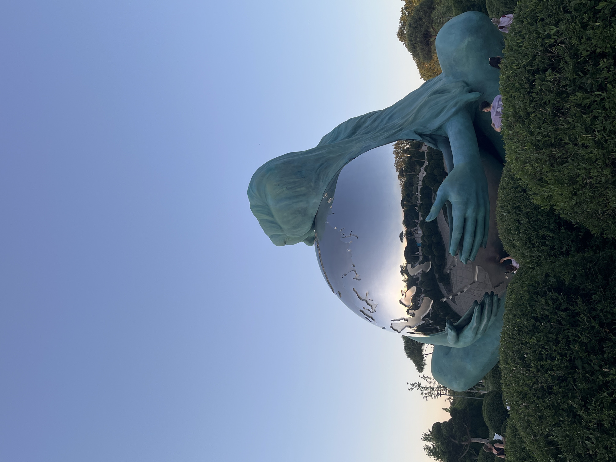 The image captures a large, teal-colored sculpture depicting a figure embracing a mirrored globe. The sculpture is situated outdoors, surrounded by manicured green bushes and trees, under a clear, pale blue sky. The mirrored globe reflects the surrounding landscape, including trees, buildings, and a sunset or sunrise with warm hues. Several people are visible in the background, some interacting with the sculpture or walking through the park-like setting. This sculpture is located in the “Galitsky Park” in Krasnodar, Russia.