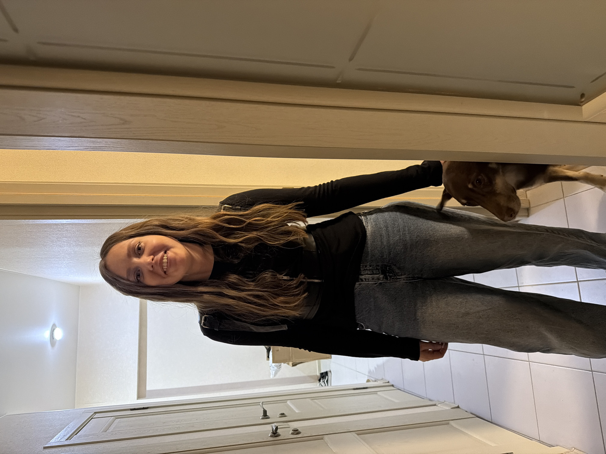 A young woman wearing jeans and a black long-sleeved shirt stands in a hallway with white tiled floors and white paneled doors. The woman is smiling and facing the camera. A brown dog is partially visible near her right arm, seemingly behind a partially obscured object. The location is not discernable from the provided image. The city of Moscow, Russia, cannot be inferred from the image.