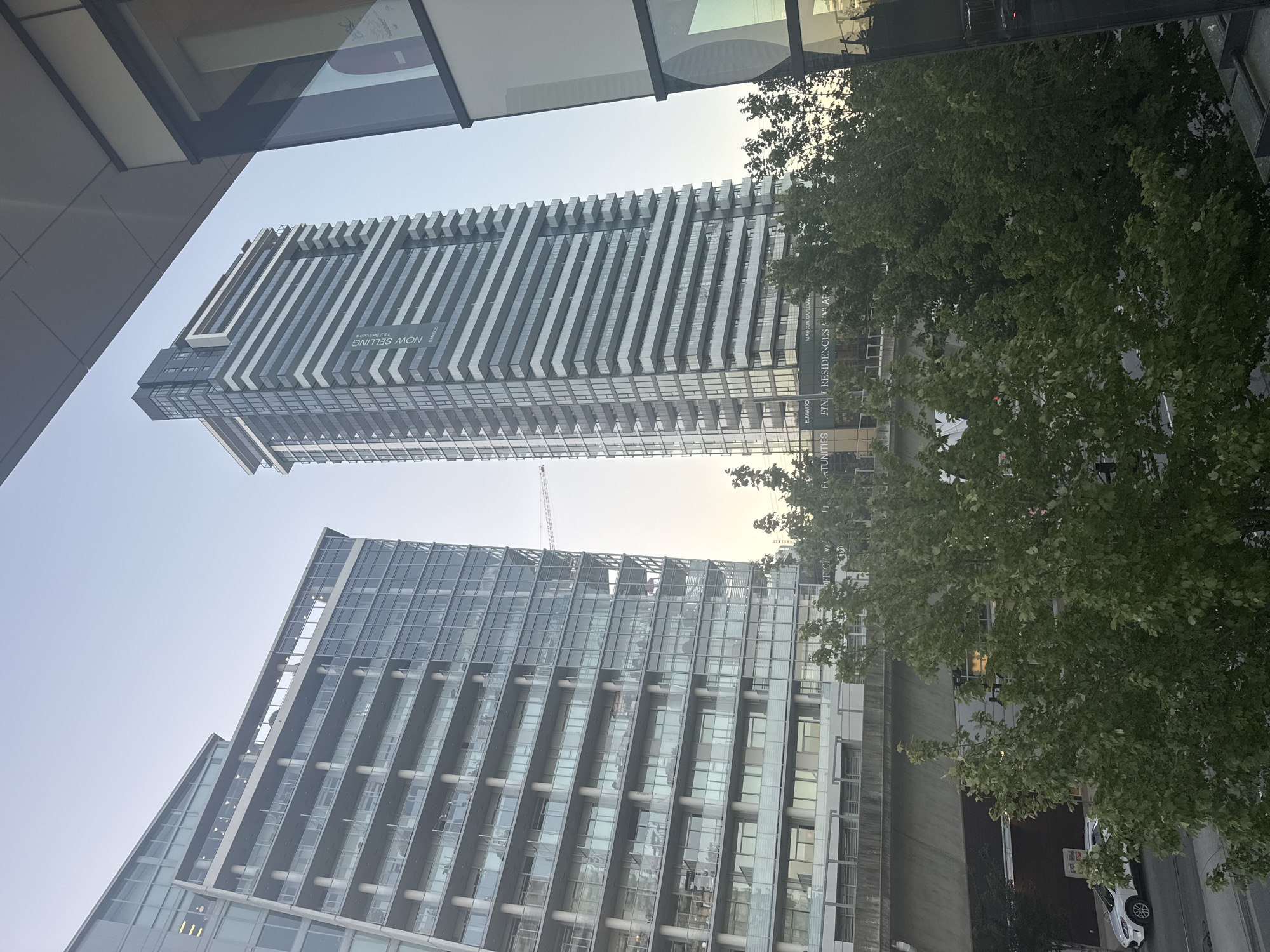 The image shows a view of modern high-rise buildings and trees in Coquitlam, Canada. In the foreground, there are lush green trees with visible foliage. Behind the trees, a multi-story concrete structure forms an elevated platform or bridge, with a dark lower section. Beyond this, two distinct skyscrapers dominate the scene. The skyscraper on the left is wider and appears to be a residential building with numerous balconies and glass-paneled floors, featuring a stepped architectural design. The skyscraper on the right is taller and slimmer, characterized by a repeating pattern of horizontal balconies and vertical window sections, with a banner that reads "NOW SELLING" and "ELMWOOD" in green lettering. A construction crane is visible between the two skyscrapers. The sky above is clear and shows a pale blue hue, suggesting it is either early morning or late afternoon. In the lower left portion of the image, a white SUV is parked on a street. The overall impression is an urban landscape with a mix of residential and commercial architecture set against a natural element of trees.