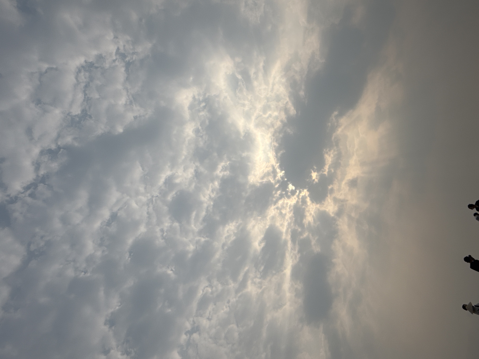 The image captures a sky with an array of clouds in Đà Nẵng, Vietnam. The clouds are predominantly gray and white, with the sun's rays breaking through in some areas, creating a bright, ethereal effect. In the lower-right corner of the frame, silhouettes of several people are partially visible. The overall environment suggests a cloudy or overcast day in an urban setting.