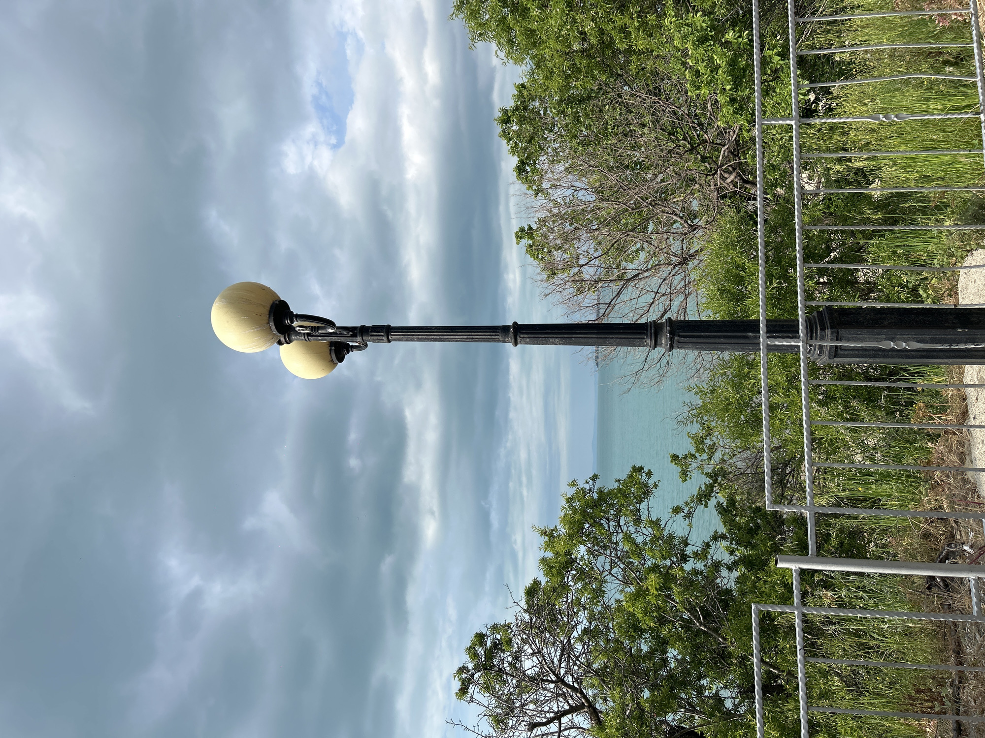 A street lamp with two spherical lights stands near a fence and greenery in Sveti Vlas, Bulgaria. The sky is overcast. The Black Sea is visible in the background.