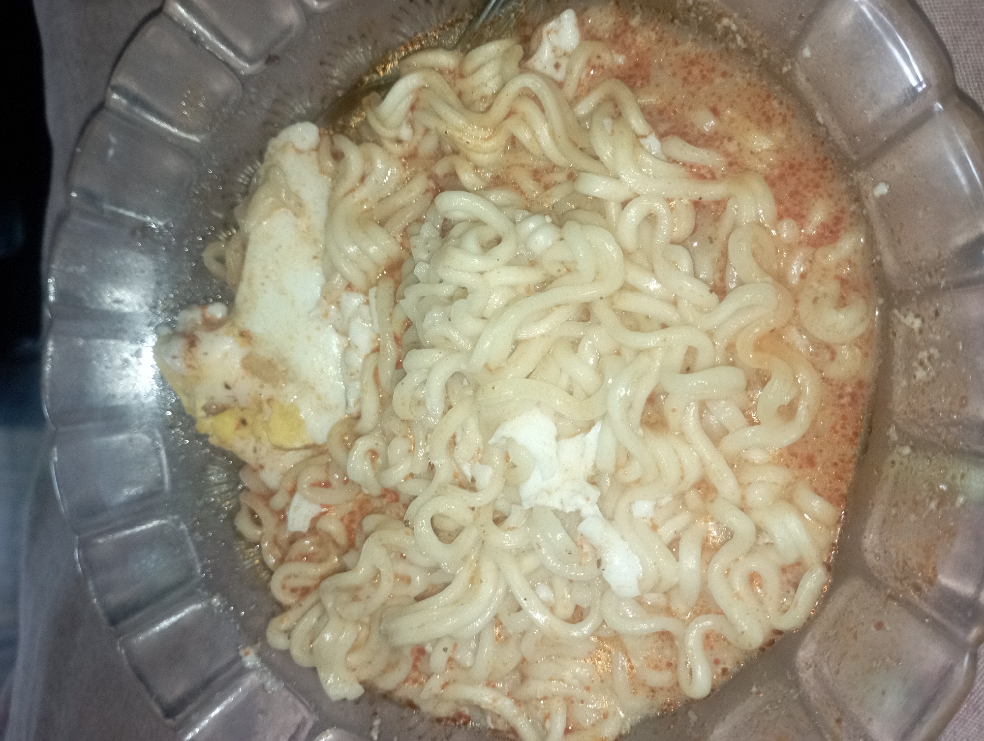 A close-up, overhead shot shows a bowl of instant noodles with a fried egg. The noodles are coiled and covered in a light orange broth. Speckled throughout the broth are small red flakes, likely chili powder. A portion of a fried egg, with a slightly runny yolk, sits atop the noodles. The bowl appears to be made of clear, fluted glass, and a patterned tablecloth is visible underneath. This dish was photographed in Maos Kidul, Indonesia.