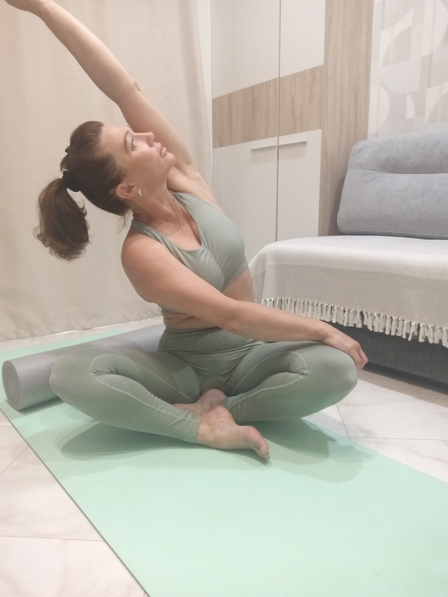 A woman with her hair in a ponytail, wearing a light green athletic outfit, is performing a yoga pose on a light green yoga mat. She is seated with her legs crossed and her right arm extended upwards and bent over her head, with her left arm resting on her left thigh. A grey foam roller is placed on the mat beside her left leg. The background shows a closet with light wood and white doors, and a grey sofa with a white throw blanket. The floor is tiled. The image is taken indoors. The scene does not contain any specific visual indicators placing it within the city of Sochi, Russia.