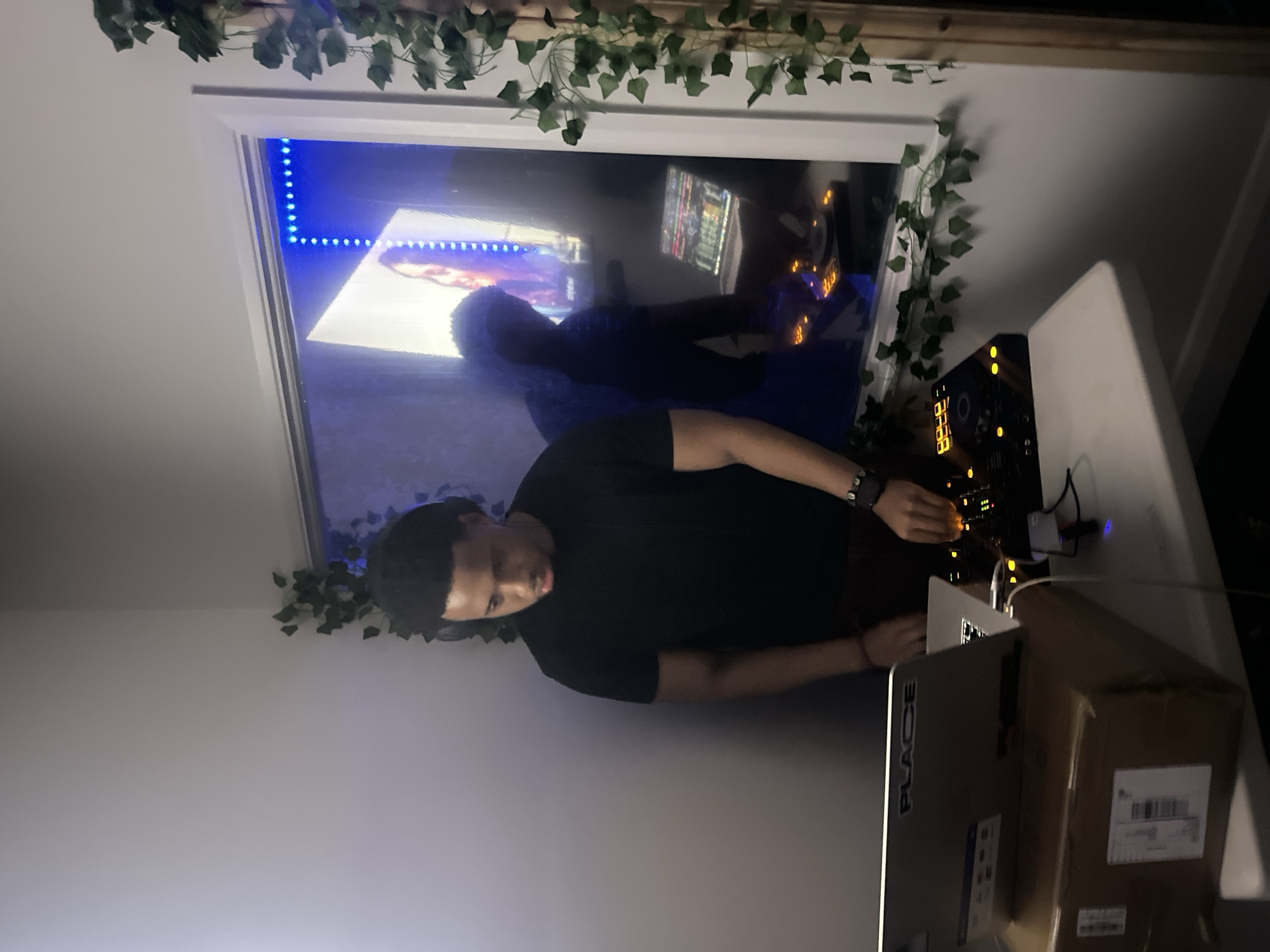A person is standing in a room, seemingly operating DJ equipment on a table. The table also contains a laptop with the word "PLACE" visible on it and a cardboard box. The room is decorated with artificial vines around a window or screen, which reflects a scene with blue lighting. No information is available to determine the location in London, United Kingdom.