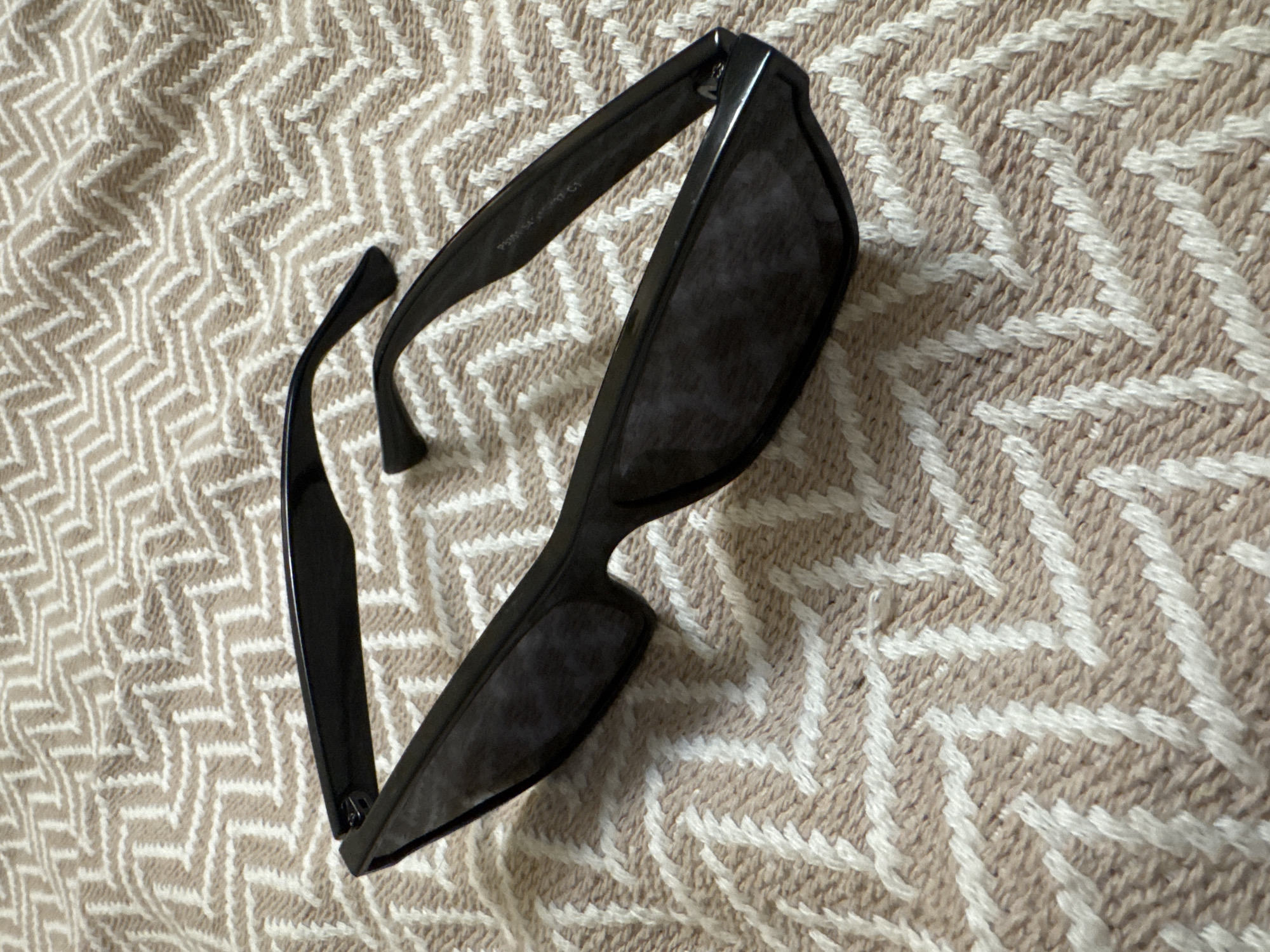 A pair of black sunglasses rests on a beige and white textured blanket. The blanket has a zig-zag pattern. The sunglasses are positioned diagonally across the frame, with the lenses facing upwards. The right arm of the sunglasses is extended, while the left arm is slightly folded. There is some text visible on the right arm of the sunglasses, but it is too blurry to read. The lighting suggests an indoor environment, possibly a bedroom or living area. There is no information available about the location in Omsk, Russia.