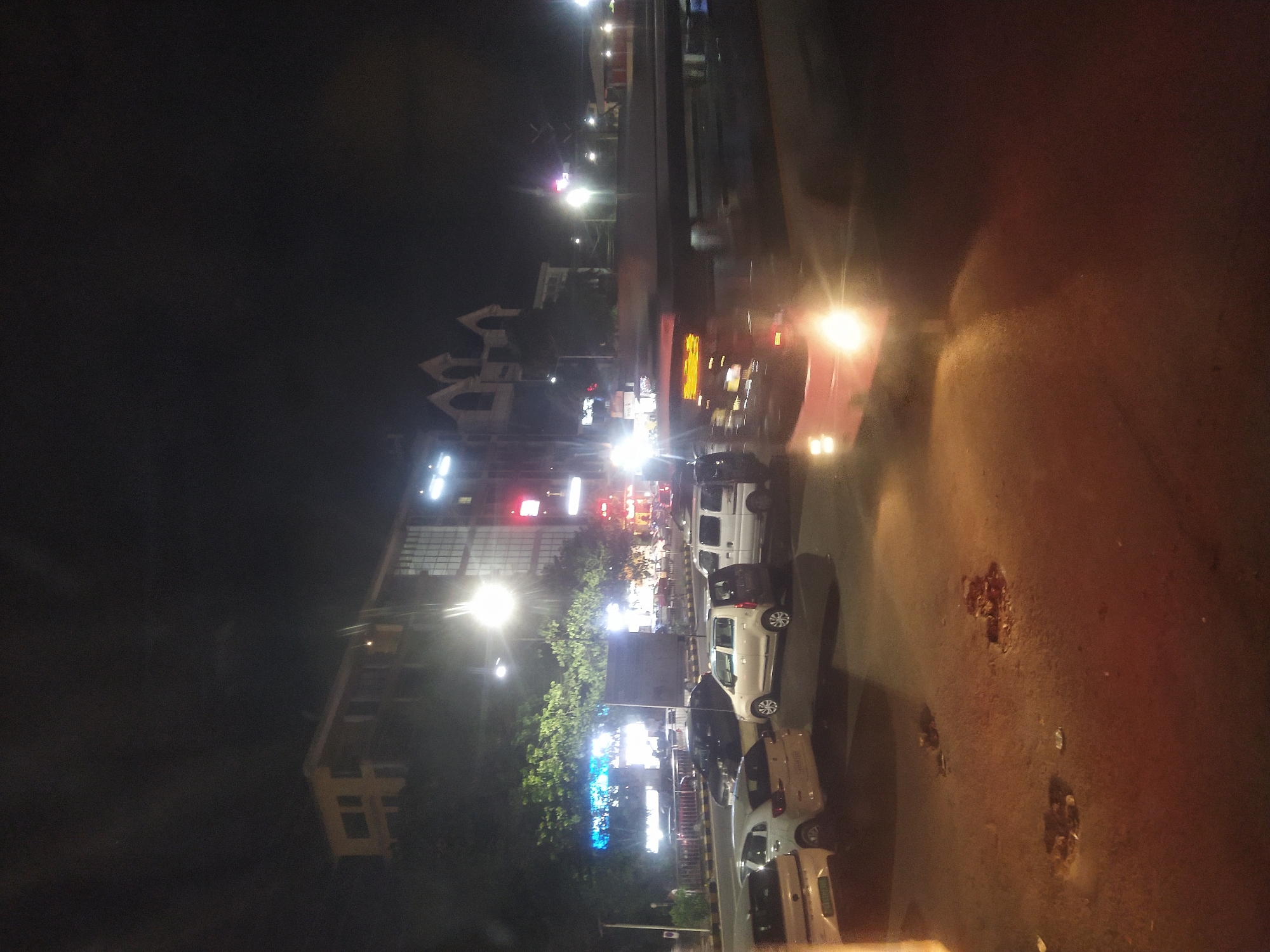 Nighttime street scene in Margao, India. Cars are parked along the side of the road. Streetlights and building lights illuminate the area. Buildings and trees line the opposite side of the road. Pavement is visible in the foreground, with some debris on the surface.