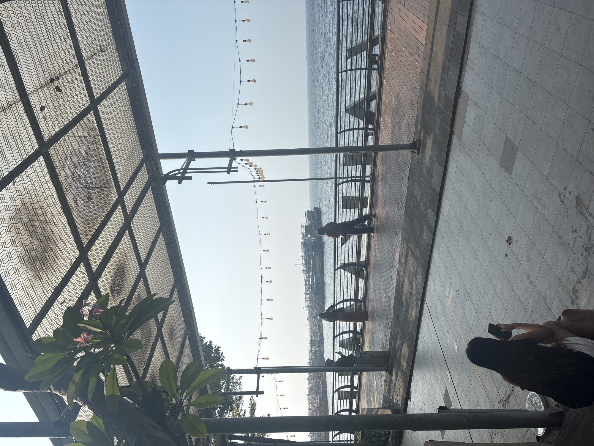 The image shows a waterfront promenade in North Jakarta, Indonesia. In the foreground, a young woman with long dark hair is sitting on a low step, looking at her smartphone. The promenade is paved with square tiles and has a tiled walkway. Beyond the walkway is a railing overlooking the sea, where several boats are docked in the distance. A string of Edison bulbs hangs overhead, casting a warm glow on the scene. The sky is clear and blue, suggesting a bright, sunny day. The architecture of the promenade includes a covered walkway with a patterned metal roof, supported by pillars. A tree with green leaves and a few pink flowers grows near one of the pillars. The overall atmosphere is relaxed and peaceful, with a few people enjoying the waterfront view.