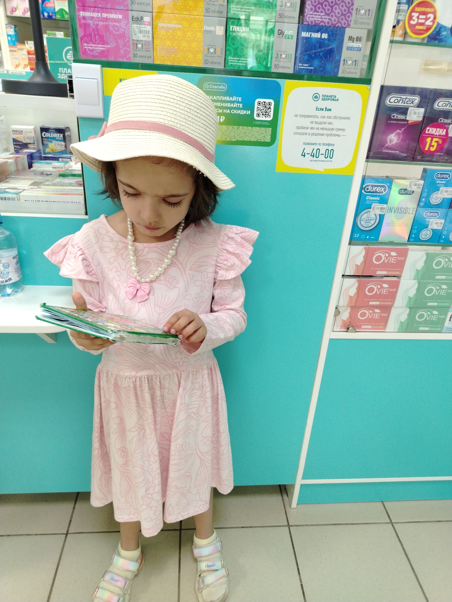 A girl stands in front of pharmacy shelves, looking at a book she holds in her hands. She wears a pink dress with ruffles at the shoulders and hem, a pearl necklace, a white hat with a pink band, socks, and colorful sandals. Behind her are shelves with pharmaceutical products, including boxes of Echinacea Premium, Eleutherococcus, Glycine, Magnesium B6, and Coldrex. Condoms and pregnancy tests are displayed to her right. A sign with a QR code and a message in Russian stating "Thank you, accumulate and exchange discounts" is visible, along with a yellow sign from the pharmacy "Planet of Health," which provides contact information in case of dissatisfaction with service at the phone number 4-40-00, located in Veltchinski, Russia.