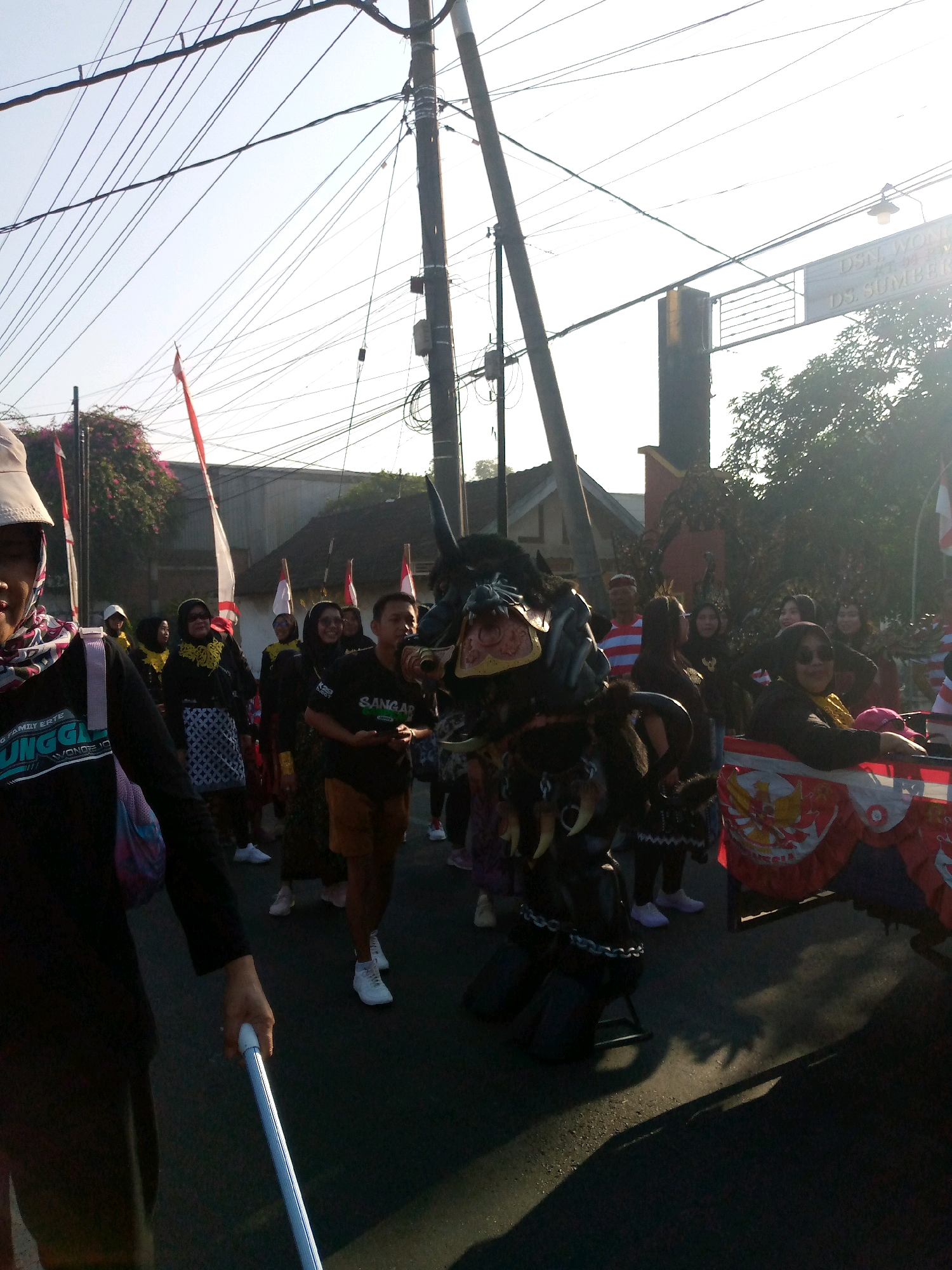 In a street scene in Sumberwono, Indonesia, a procession is underway under a bright, clear sky. The foreground is dominated by the partial view of a person in dark clothing, holding a white cylindrical object that appears to be a baton or a pole. Behind this individual, a crowd of people are participating in the event. The central focus of the procession is a large, black, animal-like costume with prominent tusks and decorative elements. This costume is adorned with chains and appears to be supported by a structure, possibly a cart or a frame, which is partially visible at the bottom.
The participants in the procession are diverse in their attire, with many wearing dark clothing, headscarves, and some carrying Indonesian flags, which are red and white. Some individuals are wearing traditional Indonesian patterned skirts. Behind the crowd, buildings and trees are visible, suggesting a residential or semi-urban environment. Electrical wires are strung across the upper portion of the image, supported by utility poles, indicating the presence of urban infrastructure. The sunlight casts long shadows on the asphalt road, suggesting it is either early morning or late afternoon. A banner with Indonesian text and the Garuda Pancasila symbol is visible on the right side of the procession, indicating a nationalistic or celebratory context. The overall atmosphere appears to be festive and community-oriented.